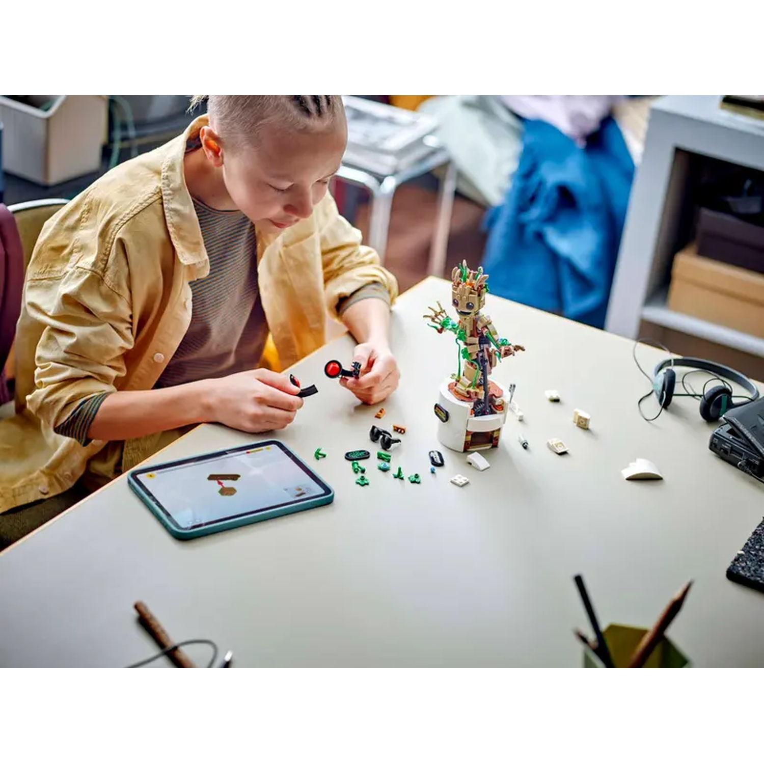 A person builds a Lego set of Groot. A tablet and various pieces are on the table. Headphones and pencils are also present.