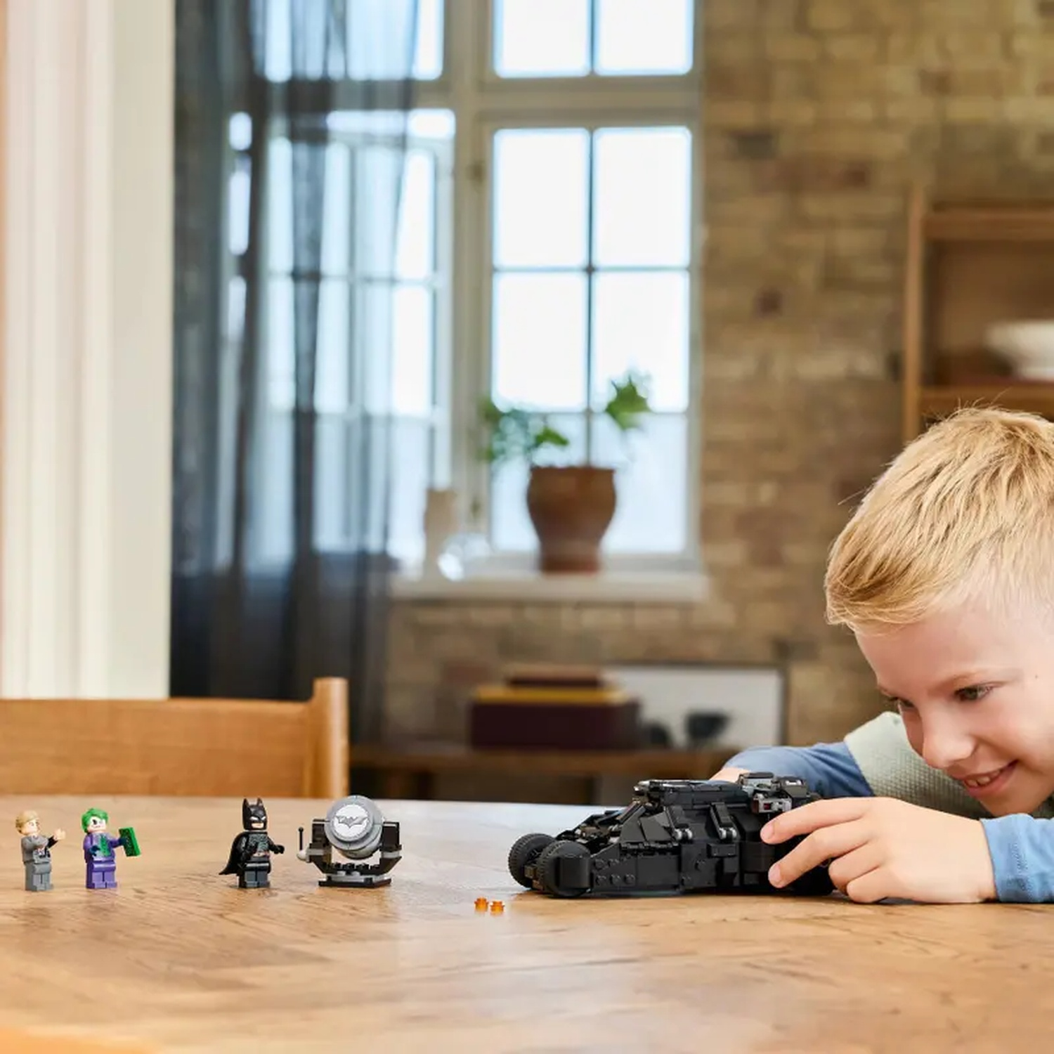Een jongen speelt met een Lego-set, inclusief Batman-figuren en een zwart voertuig, op een houten tafel.