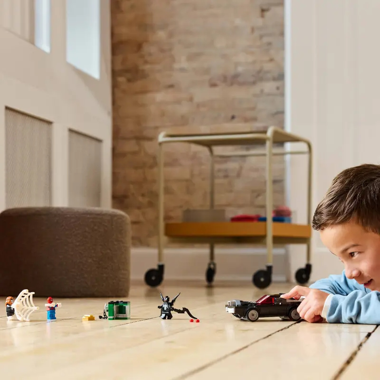A boy plays with Lego figures and a black car on a wooden floor. Various Lego characters are scattered around.