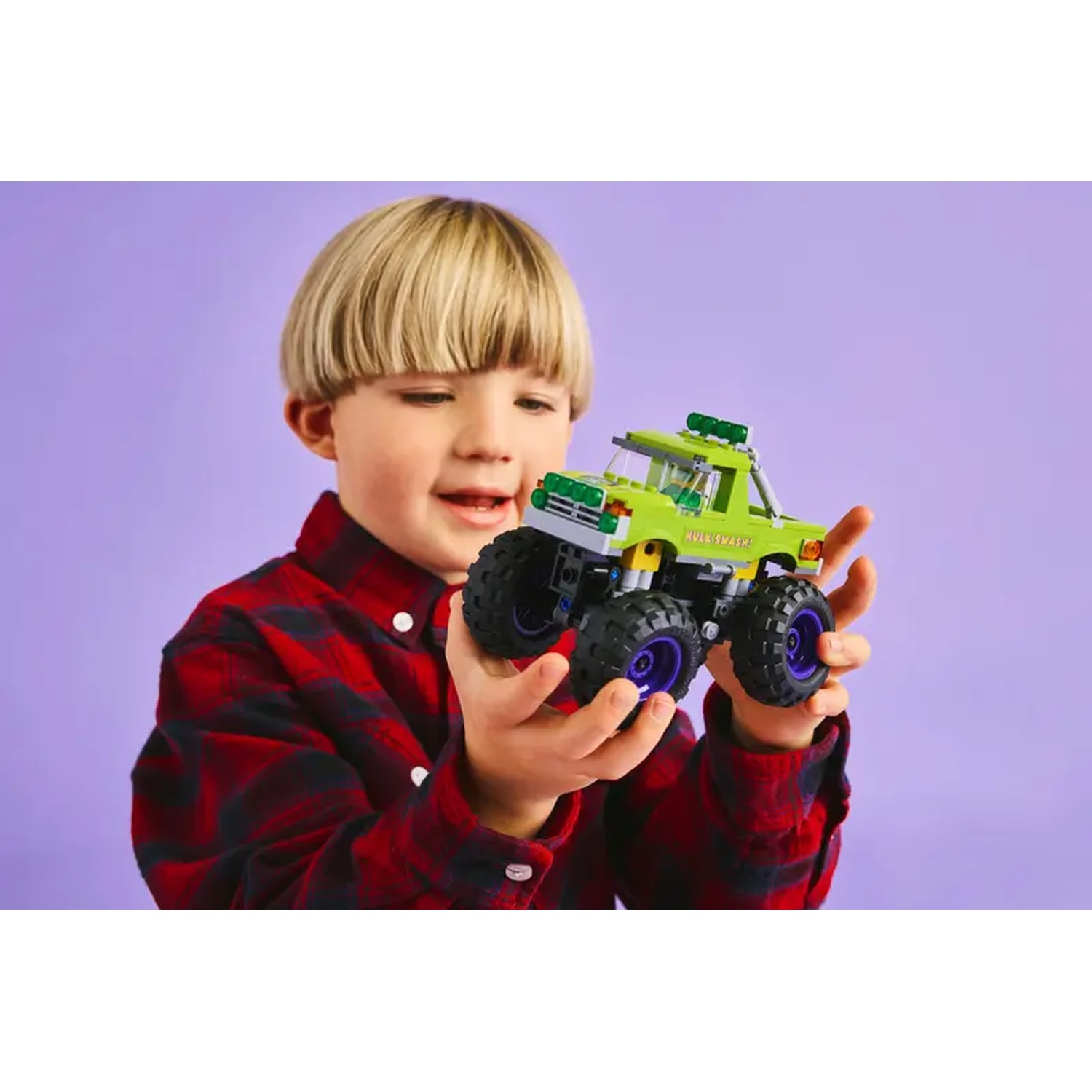 Un jeune garçon tient un camion jouet vert avec de grandes roues noires et des jantes violettes. Il porte une chemise à carreaux rouge et sourit.
