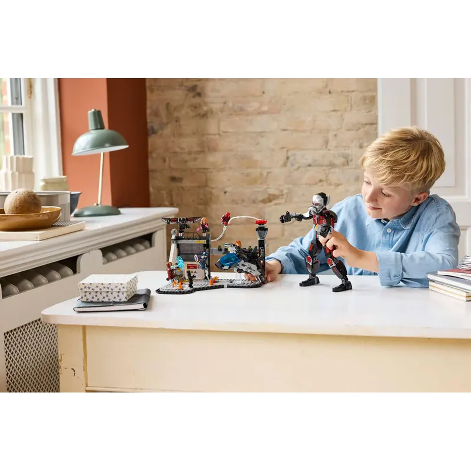 A boy plays with a Lego set, featuring action figures on a white table. A lamp and brick wall are in the background.