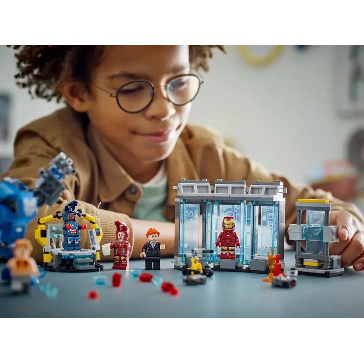 A child wearing glasses looks at a Lego set with Iron Man figures and a lab setup on a gray table.