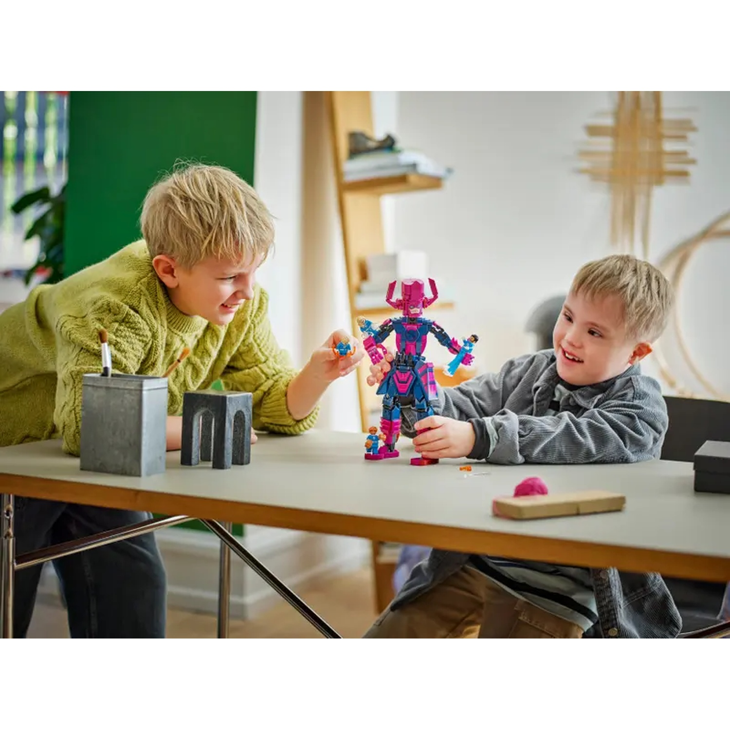 Two boys play with a Galactus Lego set. One boy holds the figure, the other reaches for a small figure. A table and objects are in view.