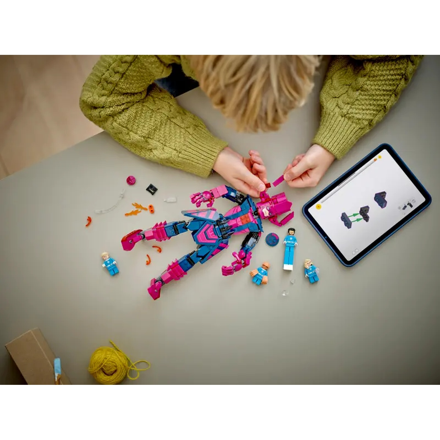 A child assembles a Lego figure with a tablet showing instructions. Lego pieces and mini-figures are scattered around.