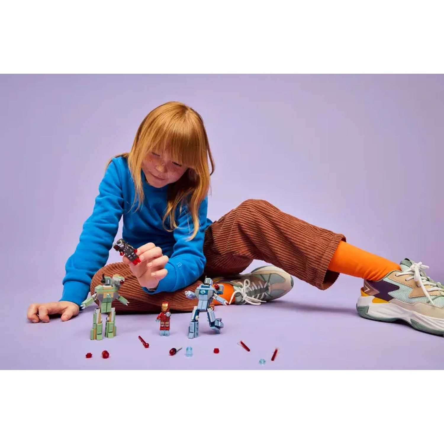 A child plays with Lego robots and figures on a purple surface. The child is wearing a blue sweater and brown pants.