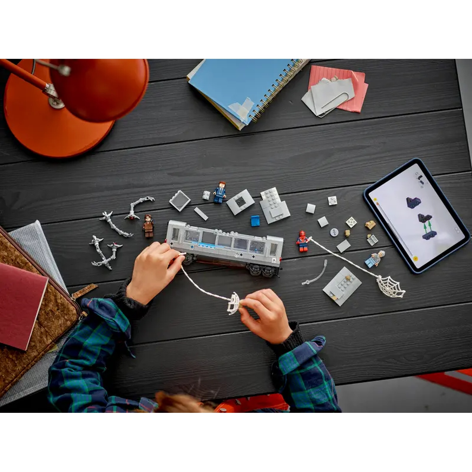 A child assembles a Lego train set on a black table, with a tablet showing instructions and various Lego pieces scattered around.