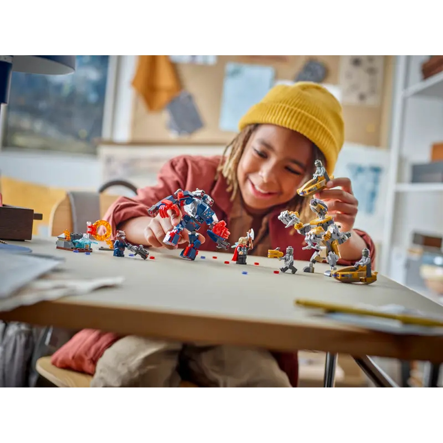 A child smiles playing with Lego toys on a table. The scene includes various Lego figures and structures with colors of red, blue, and yellow.
