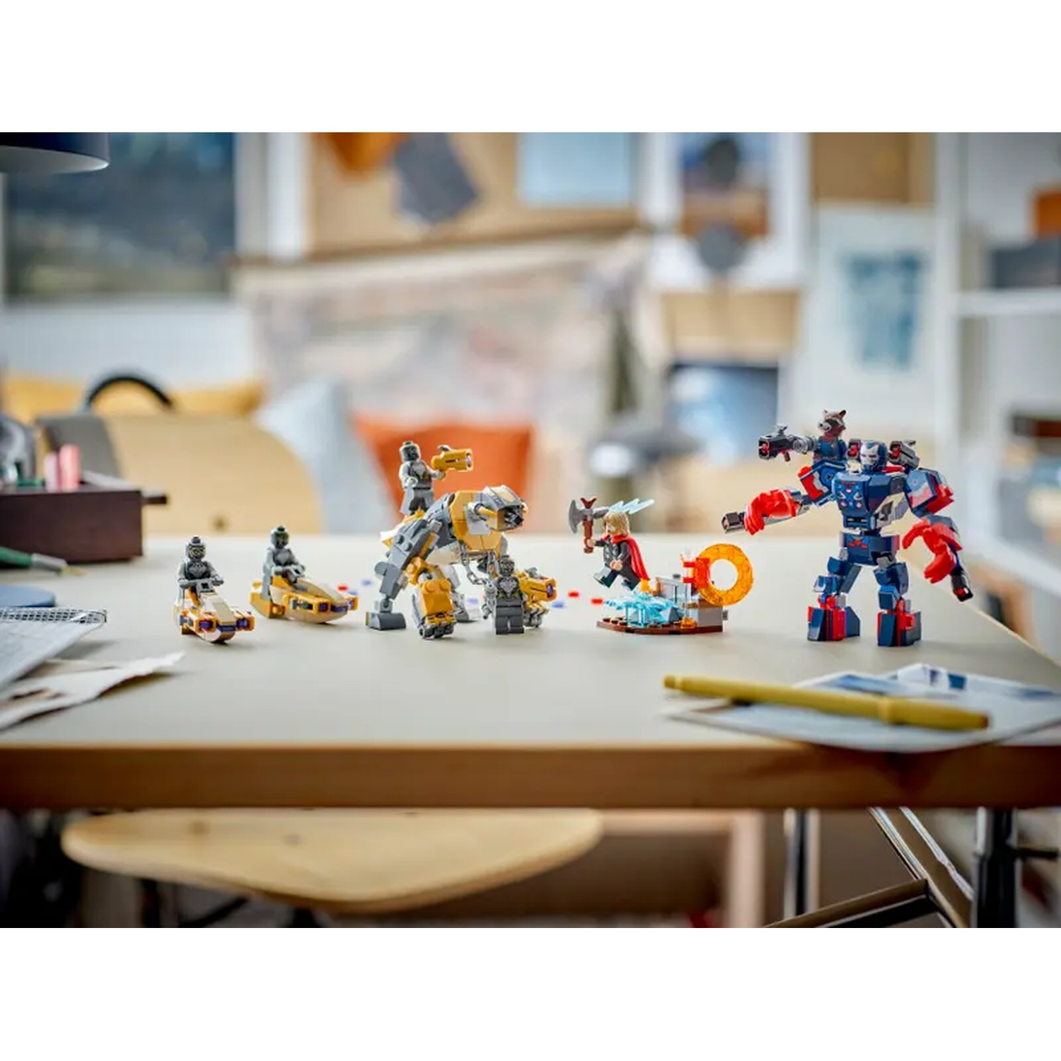 Lego sets on a table. Includes robots, characters, and vehicles. Table has papers and pen. Background has drawings and a shelf.