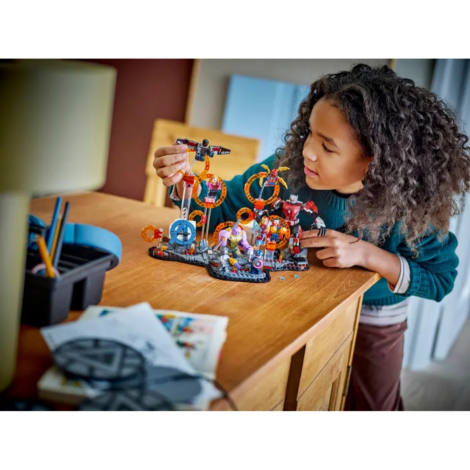 A girl with curly hair examines a Lego set on a wooden table. The set features superhero figures and circular structures.