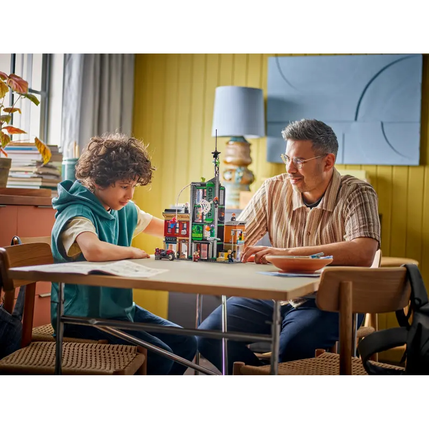 A boy and man sit at a table building a Lego set. The room is yellow. A lamp and plant are in the background.