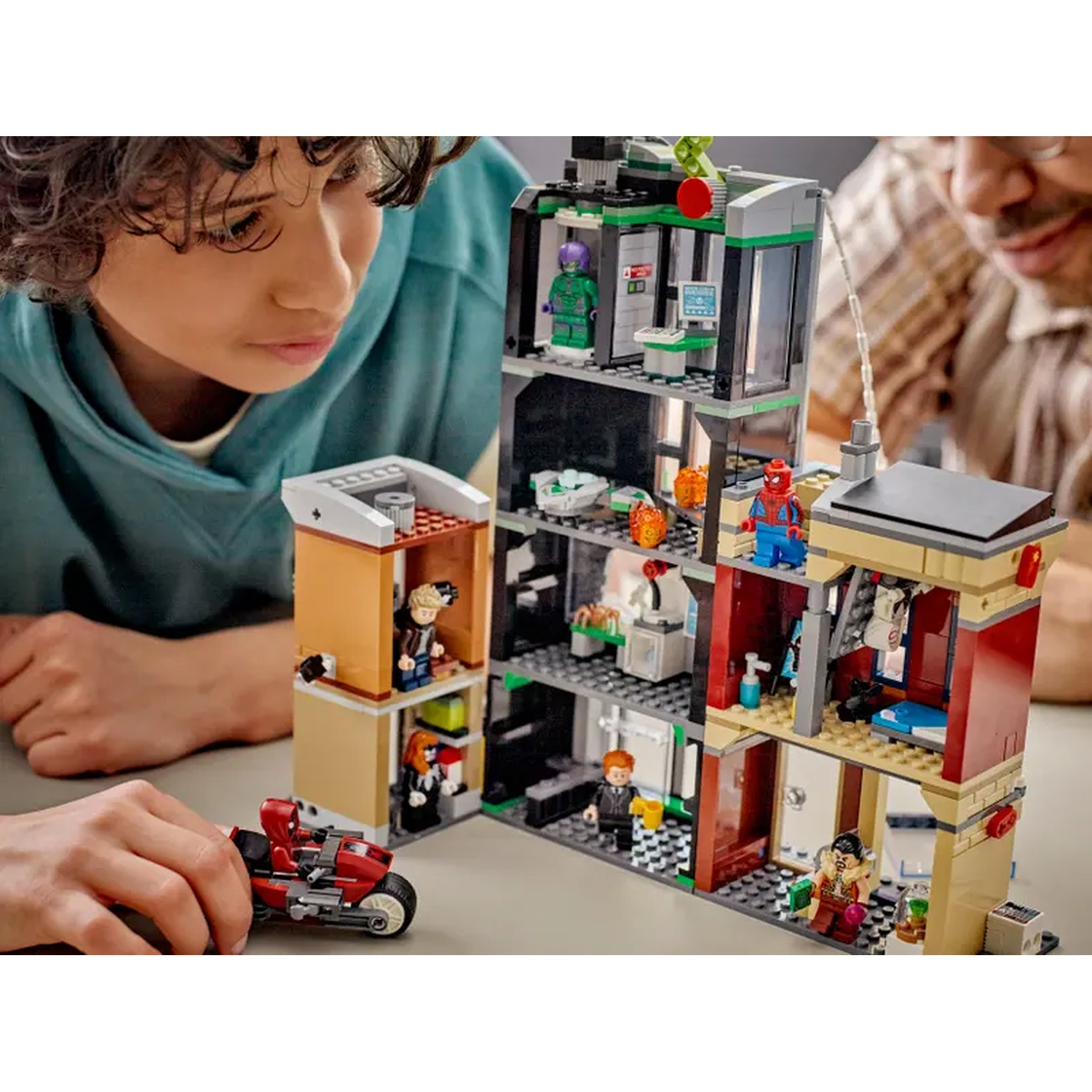 A Lego set with a building and figures. A boy and a man are looking at the set. A red motorcycle is in the front.