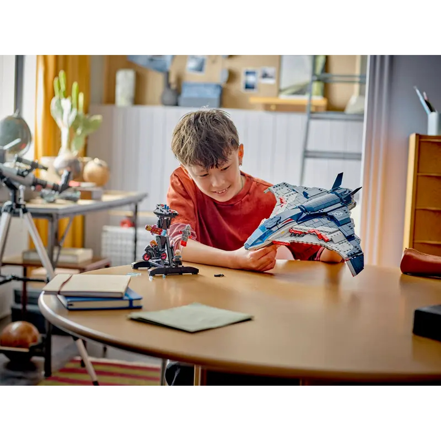 A boy smiles, holding a toy airplane. A small toy structure and books sit on a table. Cactus and a telescope are visible.