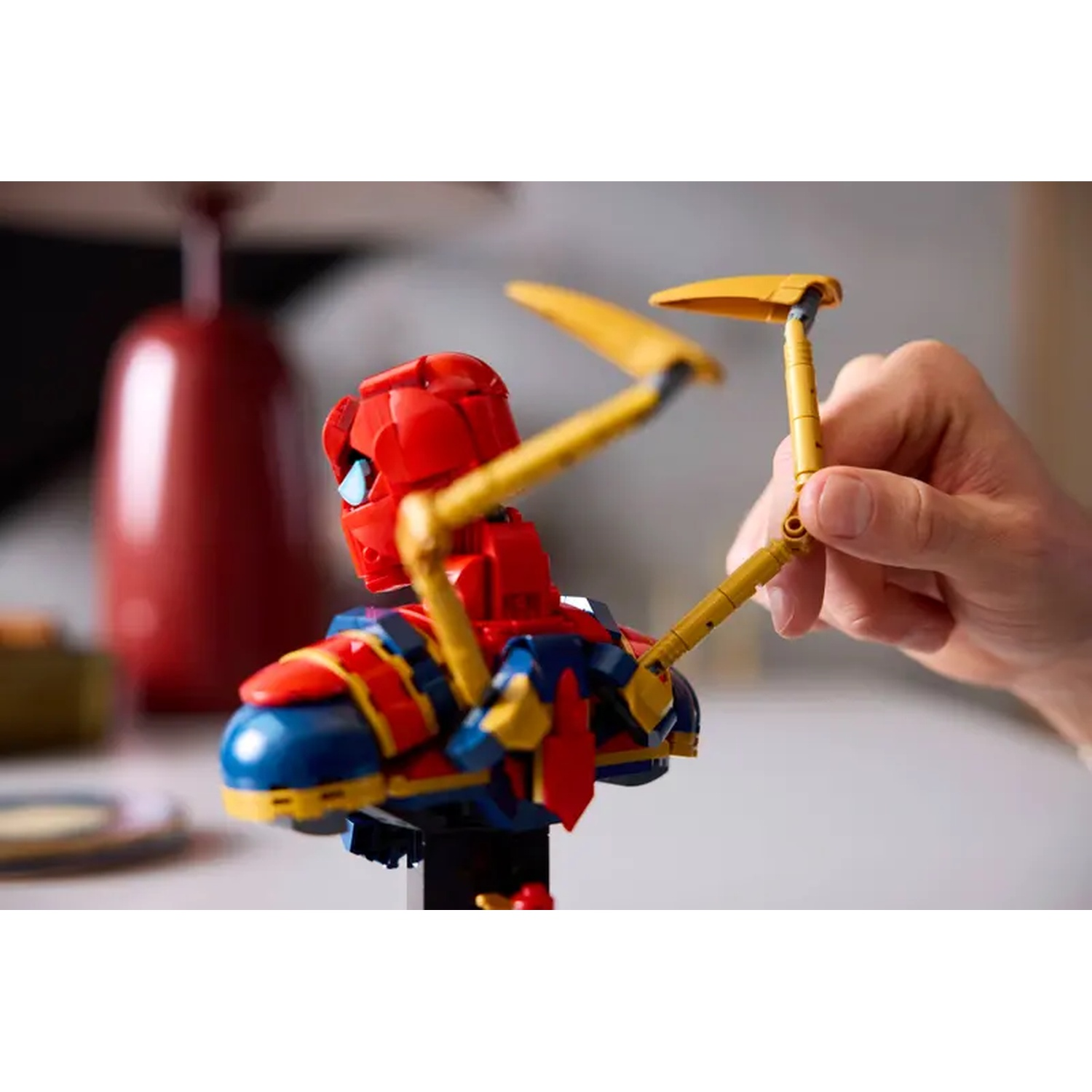 A Lego Spider-Man figure with yellow arms is held by a hand. A red vase is in the blurred background. The figure is colorful.