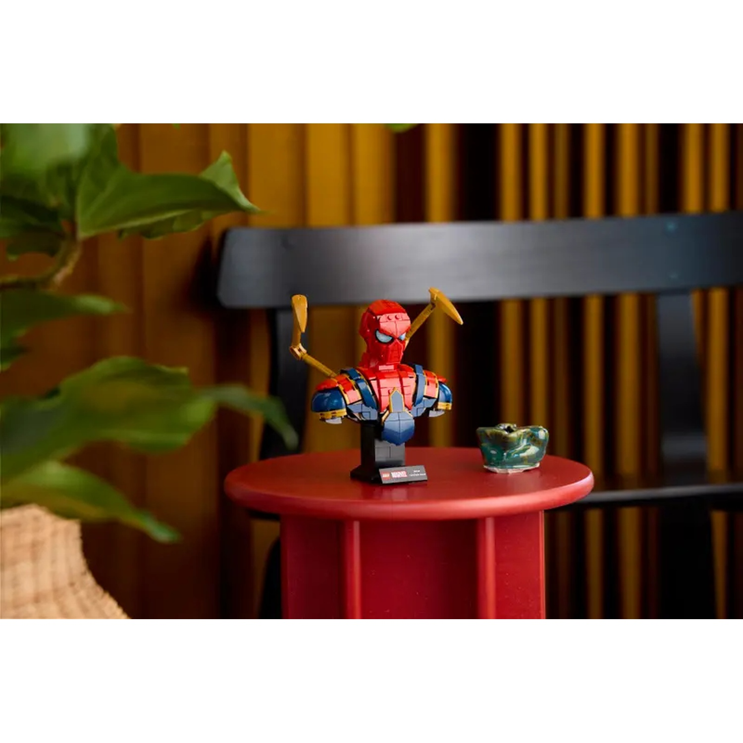 A Spider-Man bust sits on a red table, with a small green bowl. A plant is on the left, and a black bench is in the background.