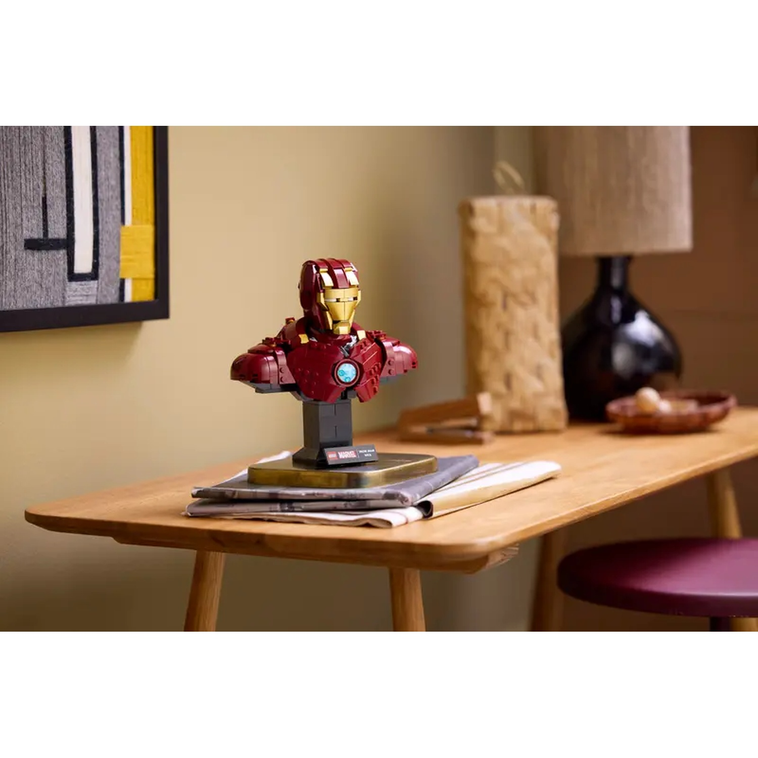 Iron Man Lego bust on a wooden table. A lamp, a stool, and other objects are in the background. A painting is on the wall.