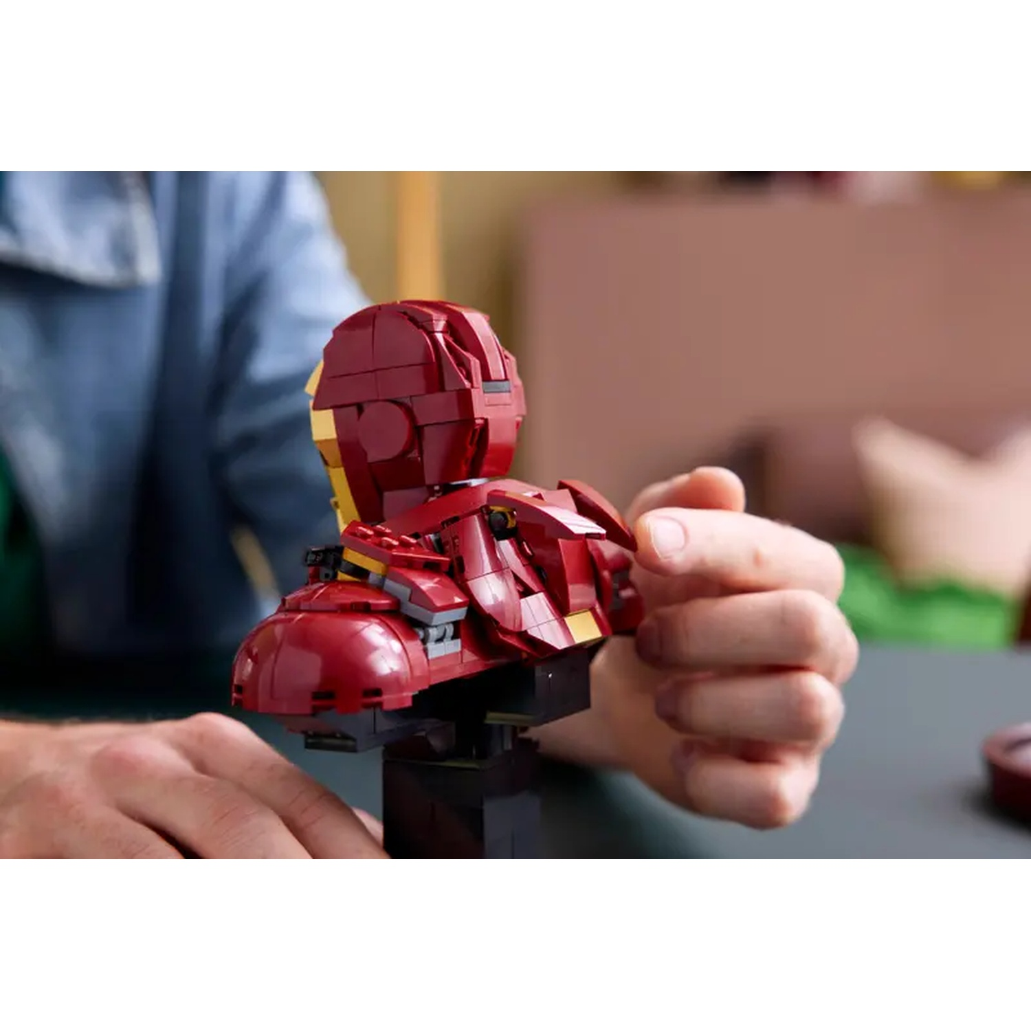 A person holding a Lego Iron Man bust. The bust is red and gold. Hands are visible. Background has green and pink hues.