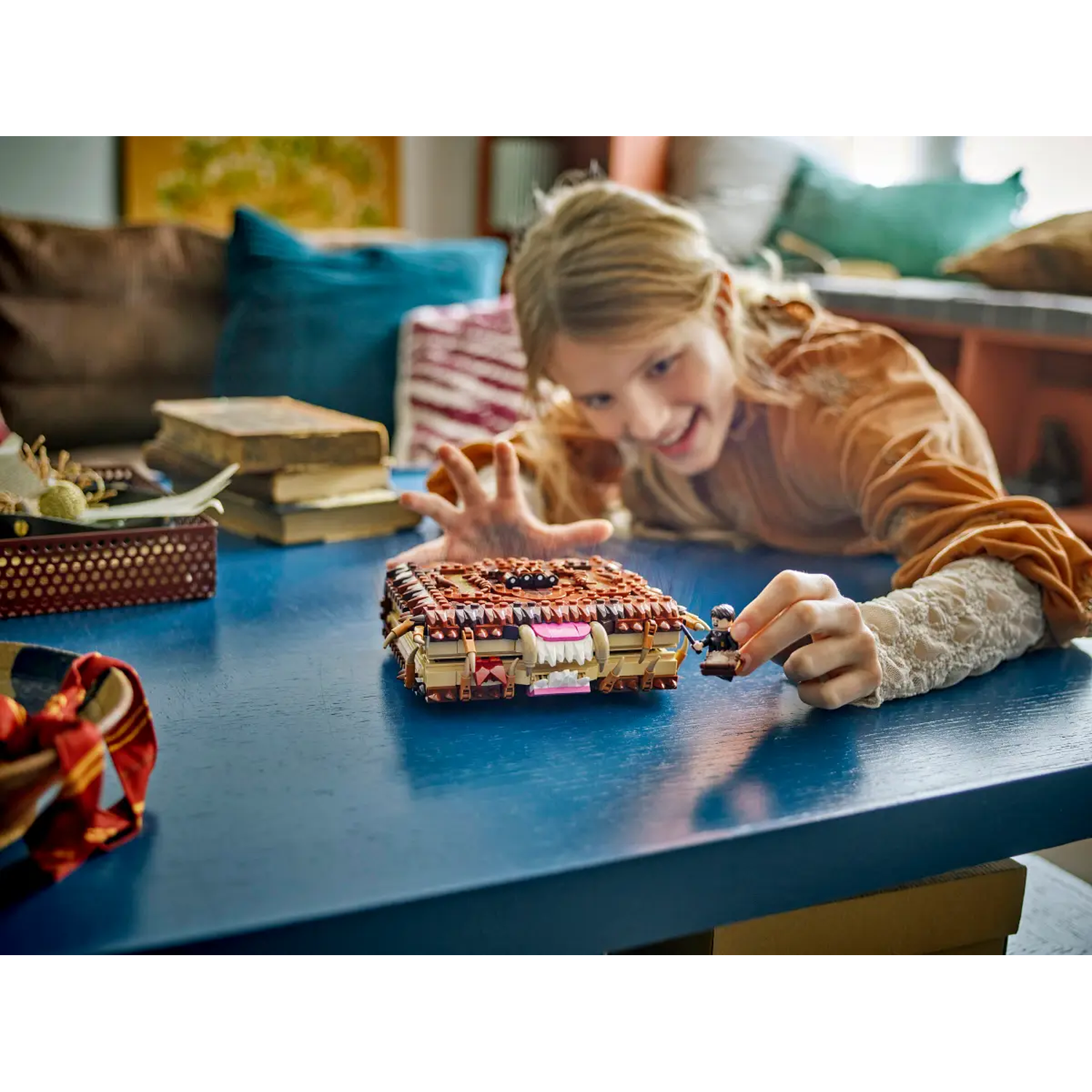 A girl plays with a Lego book on a blue table. Books and a red scarf are also on the table. The girl is smiling.