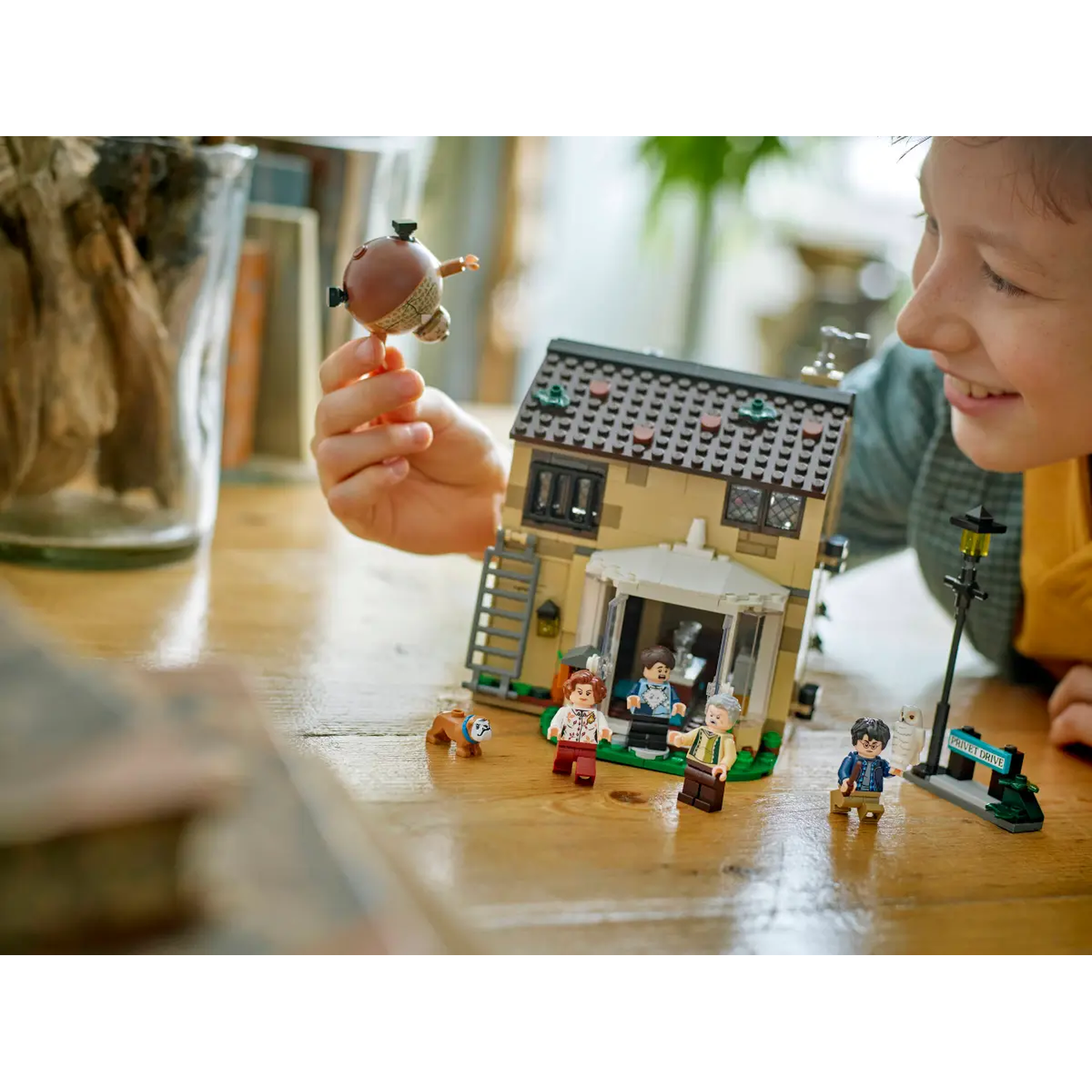 A boy holds a toy. A Lego house, figures, and a sign are on a wooden table. The boy smiles. Other objects are present.