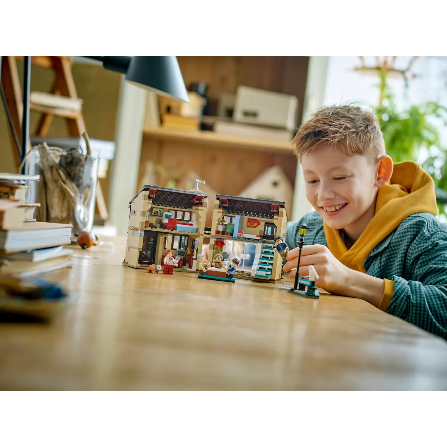 A boy smiles while playing with a Lego set of a building. He is wearing a yellow hoodie and a plaid shirt.