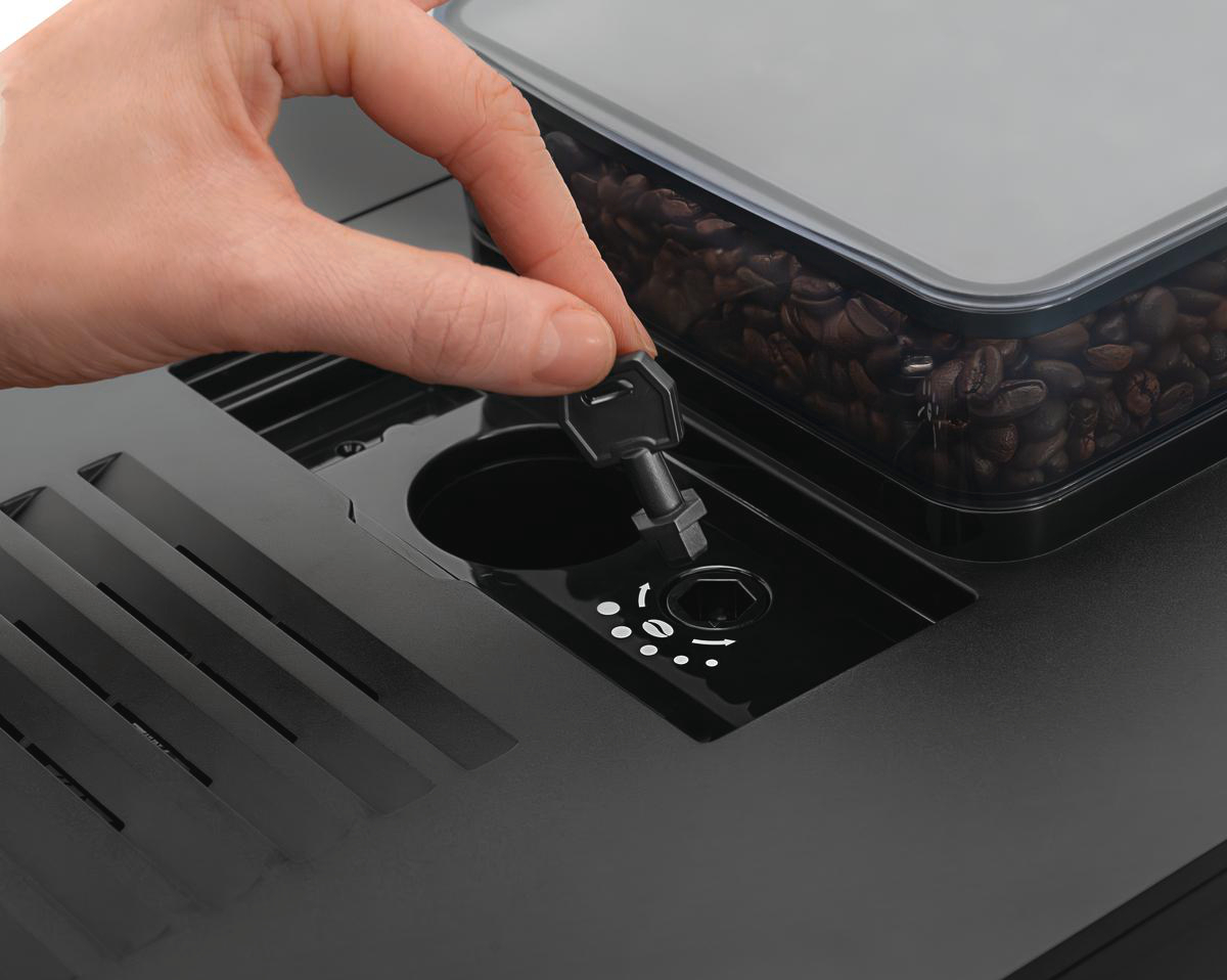 Eine Hand steckt einen Schlüssel in eine Kaffeemaschine. Ein Behälter mit Kaffeebohnen ist sichtbar. Die Maschine ist schwarz.