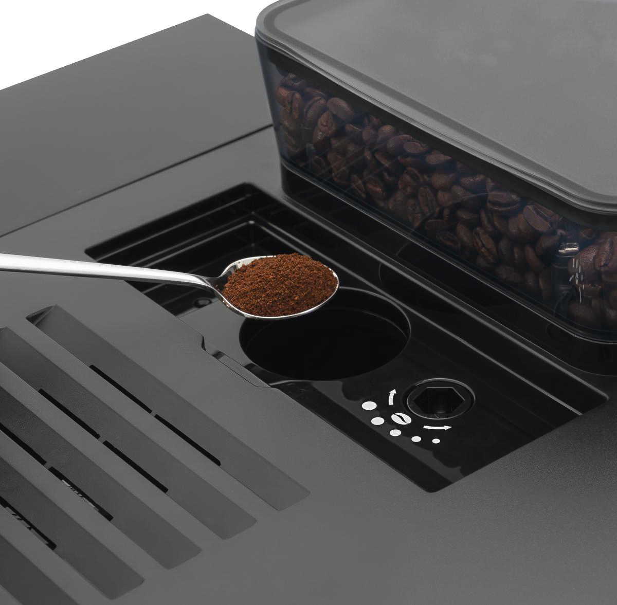 Ein Löffel hält Kaffeepulver über einer Kaffeemaschine. Ein Behälter mit Kaffeebohnen ist sichtbar. Die Maschine ist schwarz.