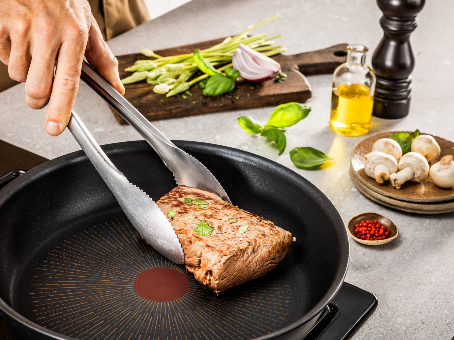 Una mano usa pinzas para voltear un bistec en una sartén. Verduras y especias están en la encimera.