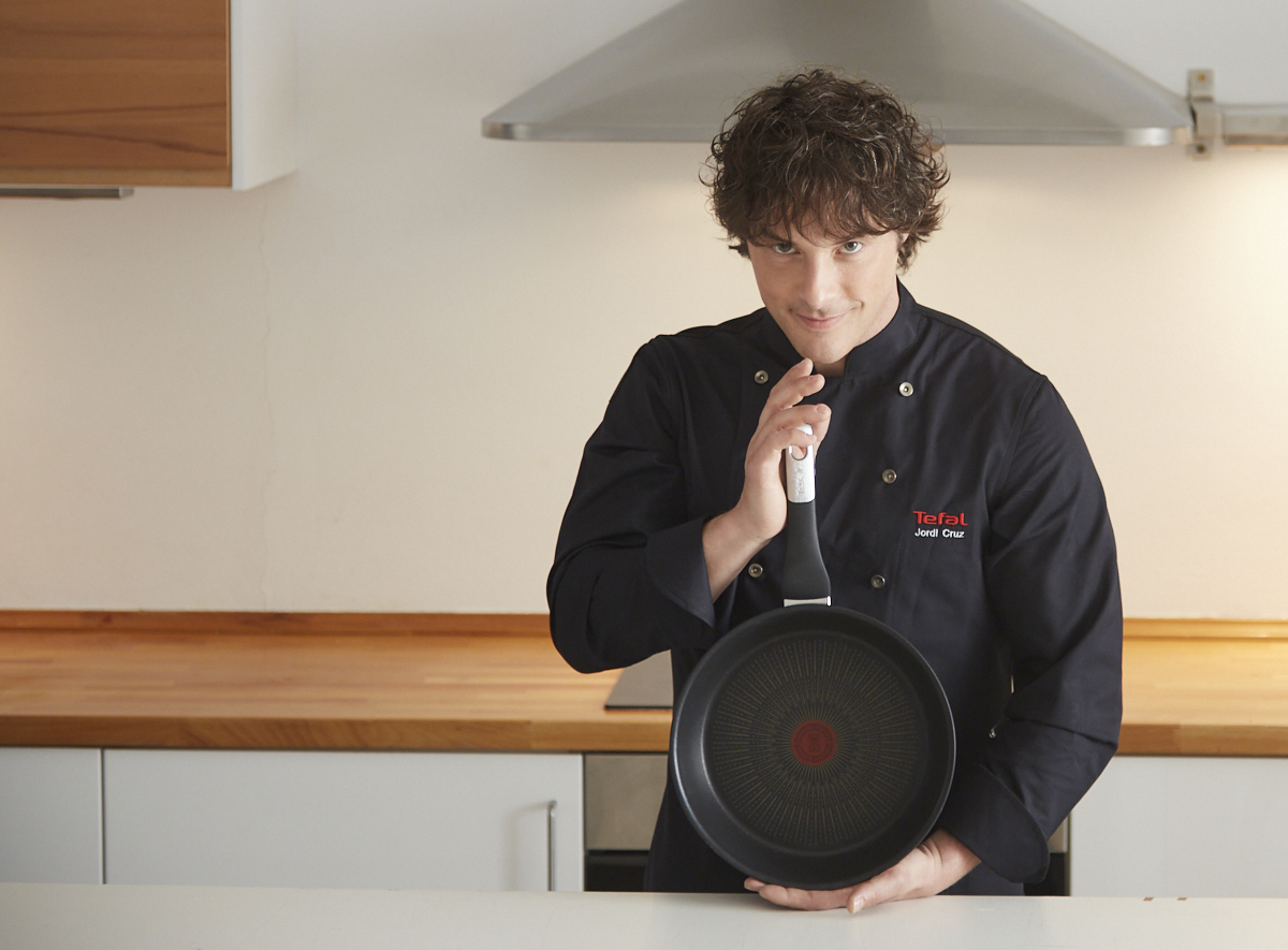 Un chef con uniforme negro sostiene una sartén Tefal, sonriendo. Ambiente de cocina con tonos blancos y madera.