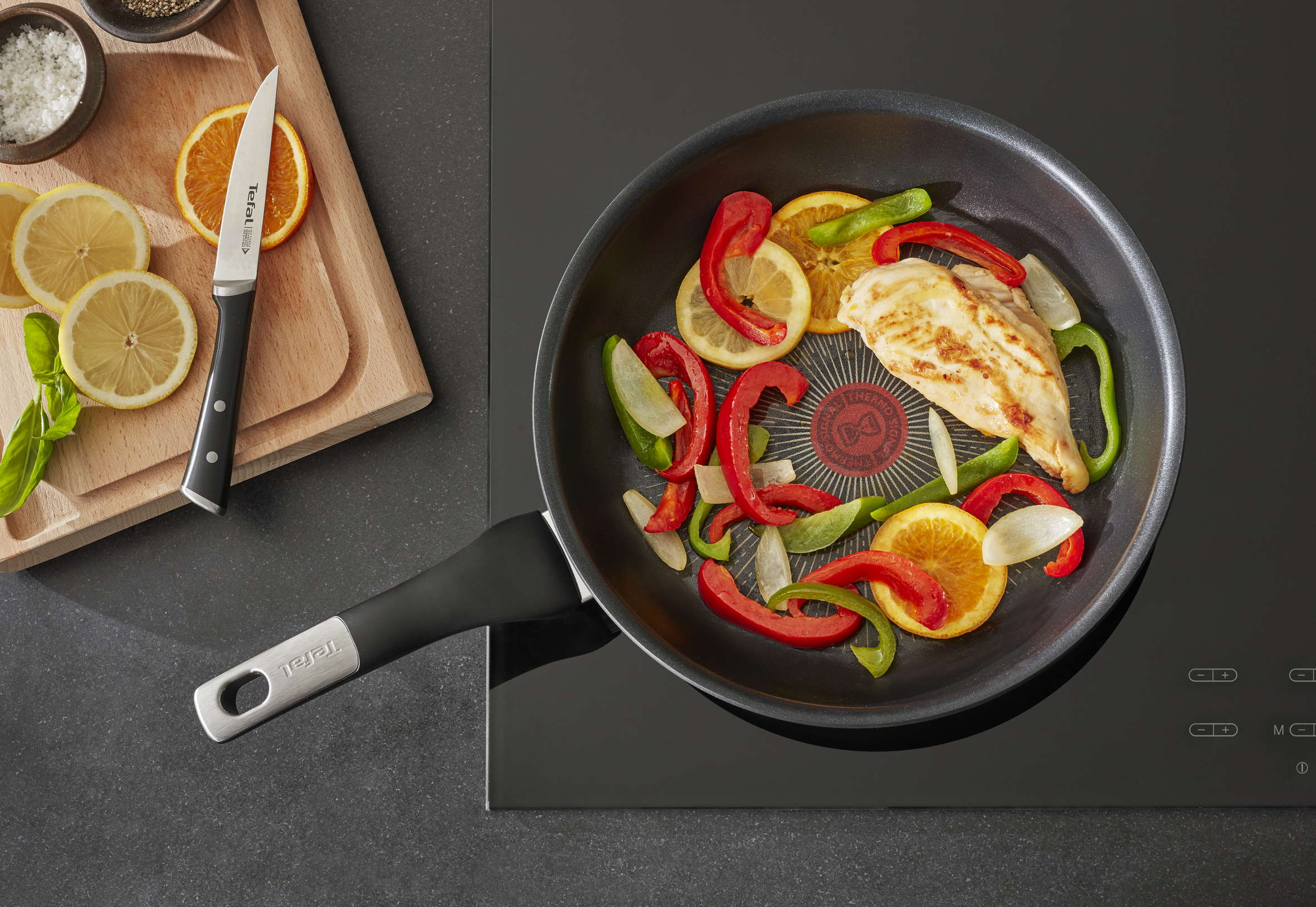 Sartén con pollo, pimientos y rodajas de naranja. Cuchillo sobre una tabla con rodajas de limón. Placa negra.