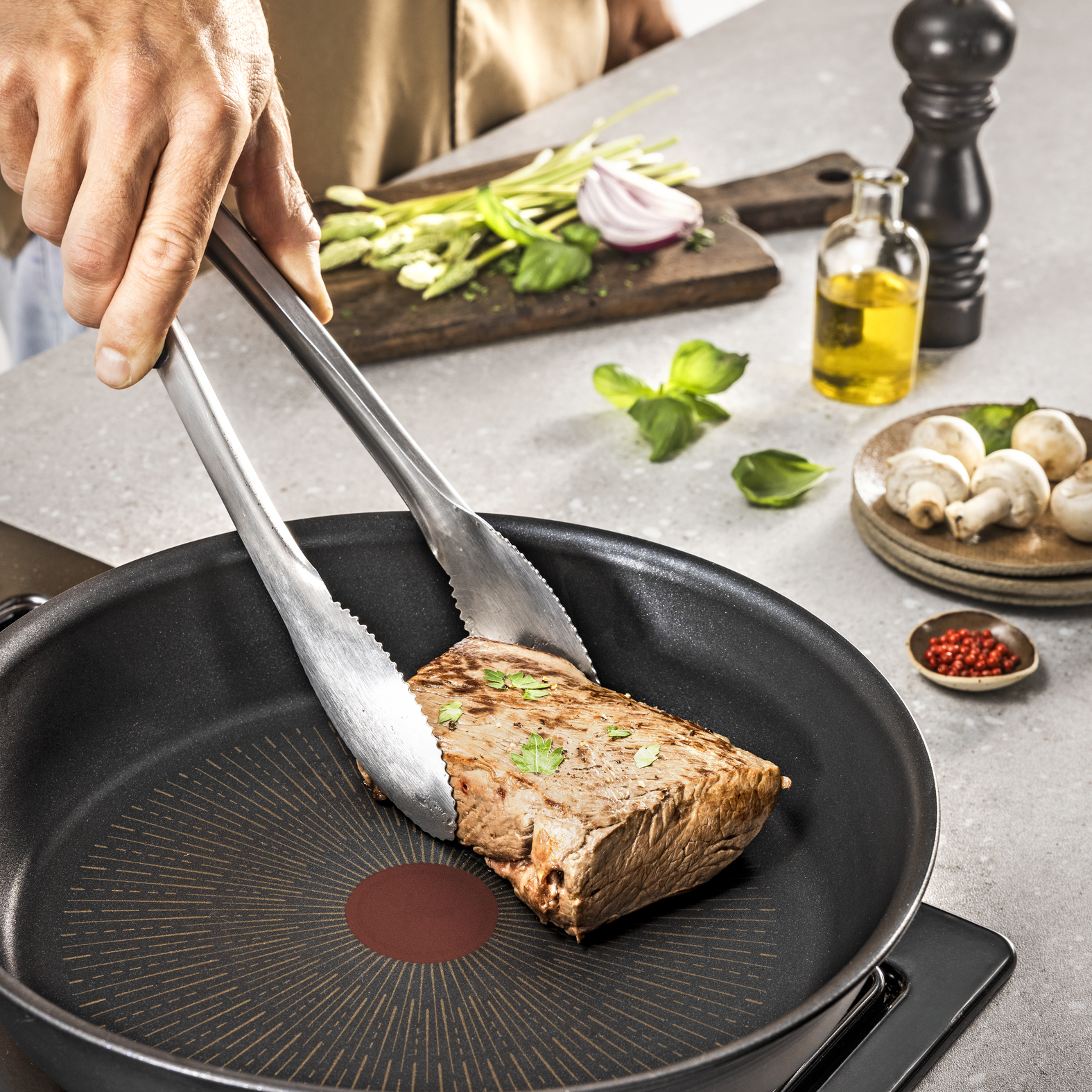 Una mano usa pinzas para voltear un bistec en una sartén. Verduras y aceite están cerca en una encimera.