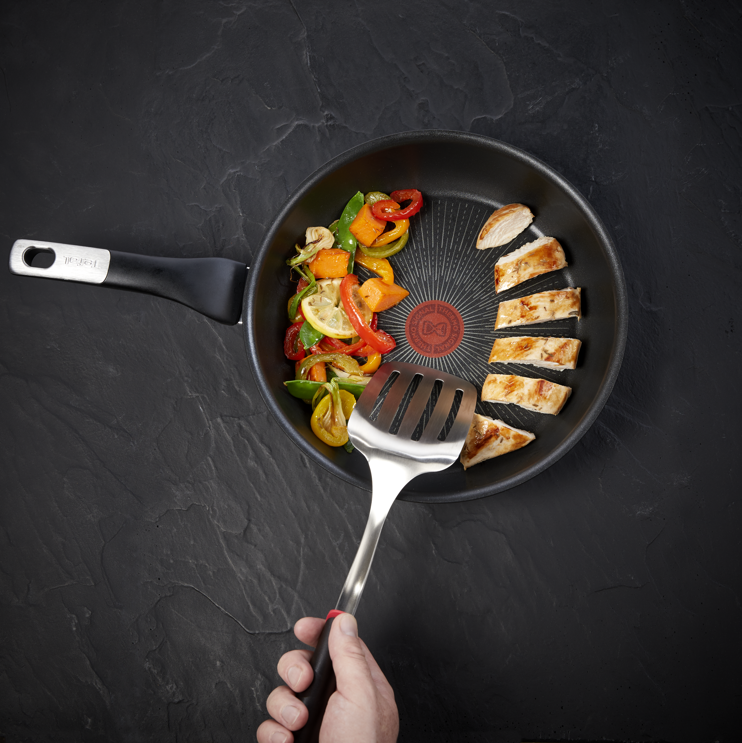 Sartén con verduras y pollo. Una mano sostiene una espátula. Fondo negro texturizado.