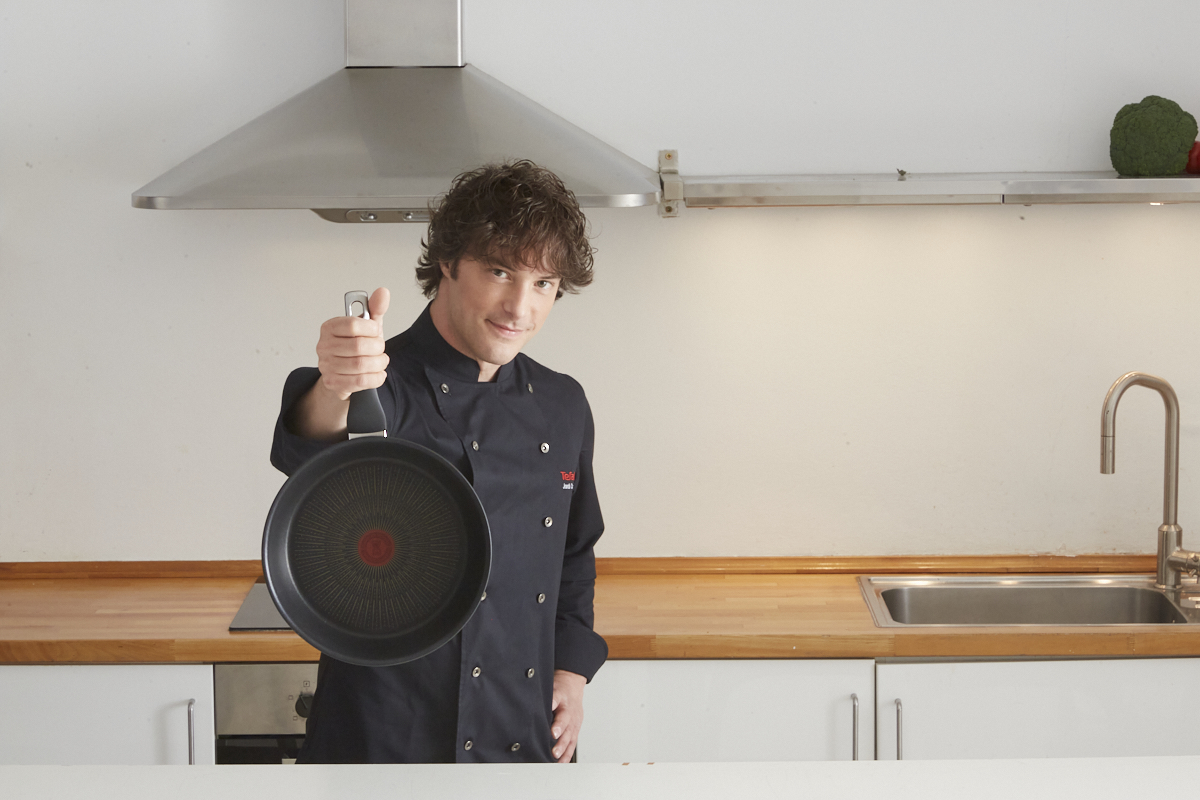 Chef con uniforme negro sostiene una sartén, sonriendo. Cocina con fregadero, campana extractora y encimera al fondo.