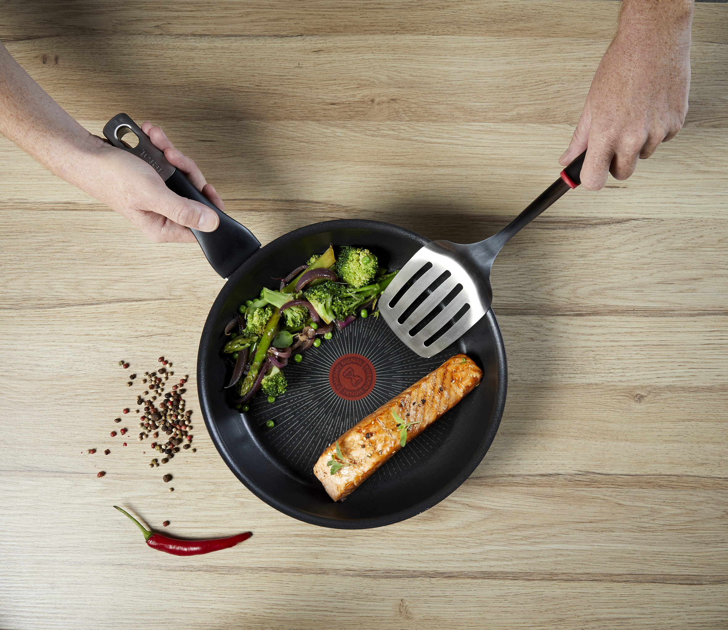 Una sartén con salmón, verduras y especias, sostenida por dos manos con una espátula sobre madera.