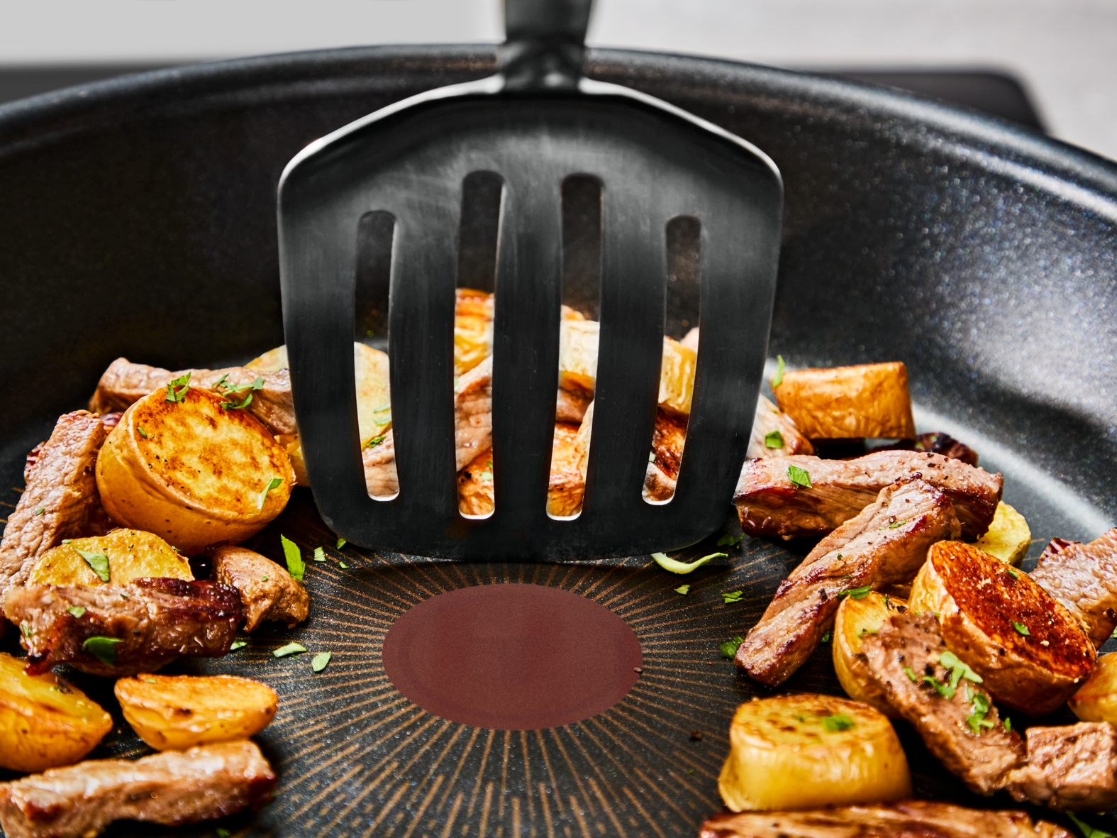 Sartén con bistec y patatas. Una espátula negra está en el centro. La carne es marrón y las patatas doradas.