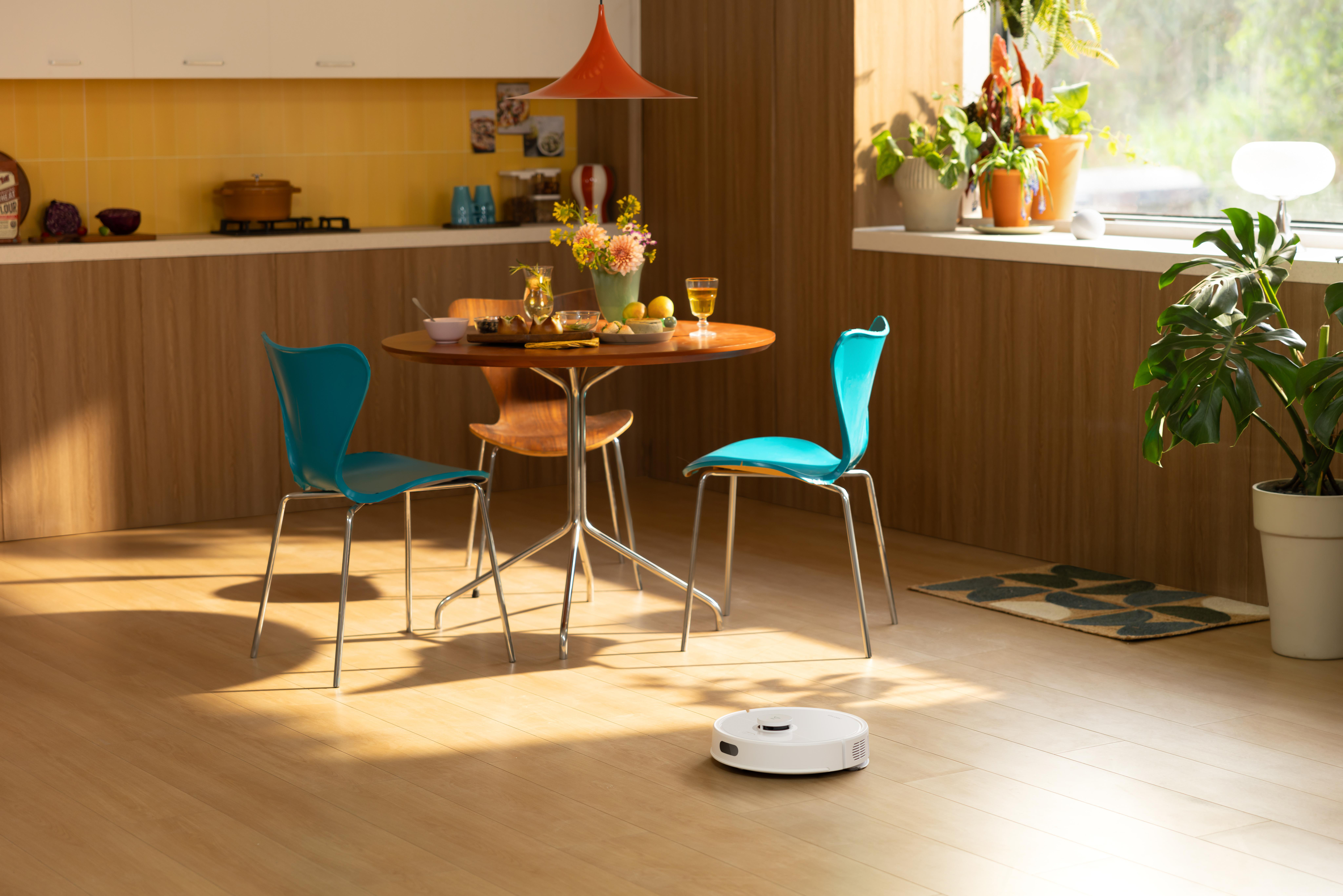 Ein Esszimmer mit Tisch, Stühlen, Blumen und einem Roboterstaubsauger auf dem Holzboden.