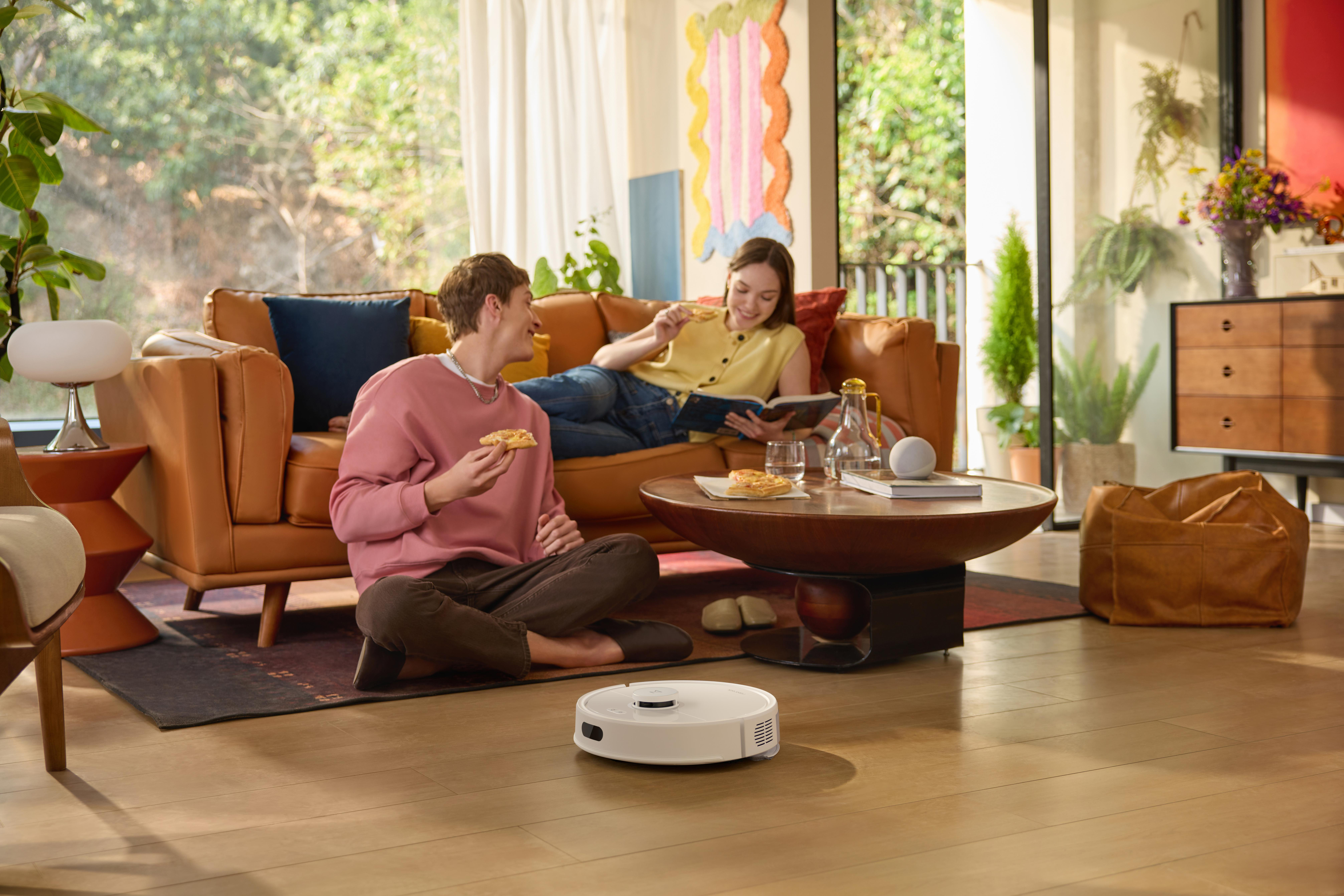 Wohnzimmerszene. Zwei Personen auf dem Sofa, eine liest. Roboter-Staubsauger auf dem Boden und ein Couchtisch.