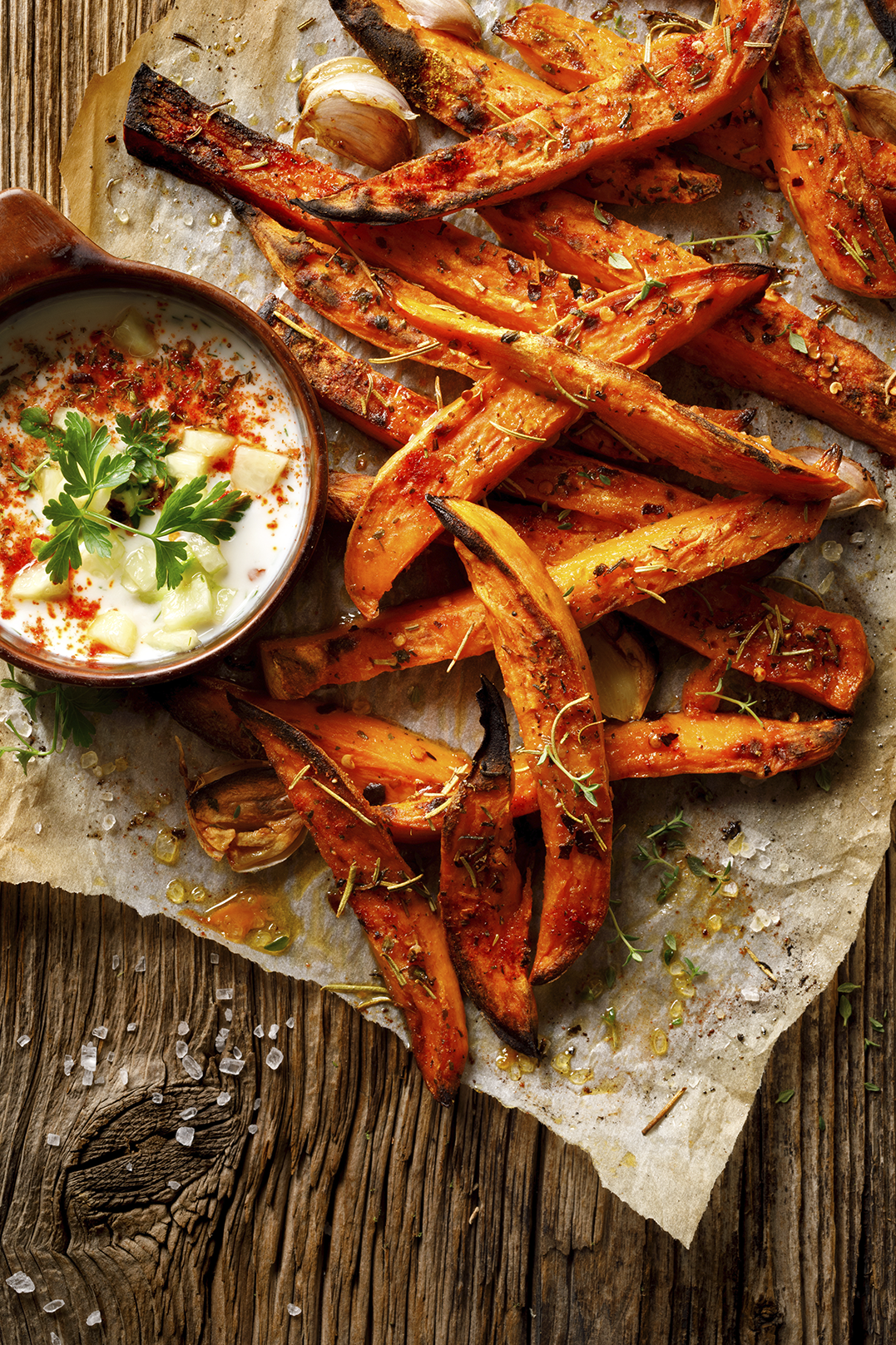 Pieczone frytki z batatów z ziołami, czosnkiem i kremowym dipem, na papierze do pieczenia na drewnianej powierzchni.