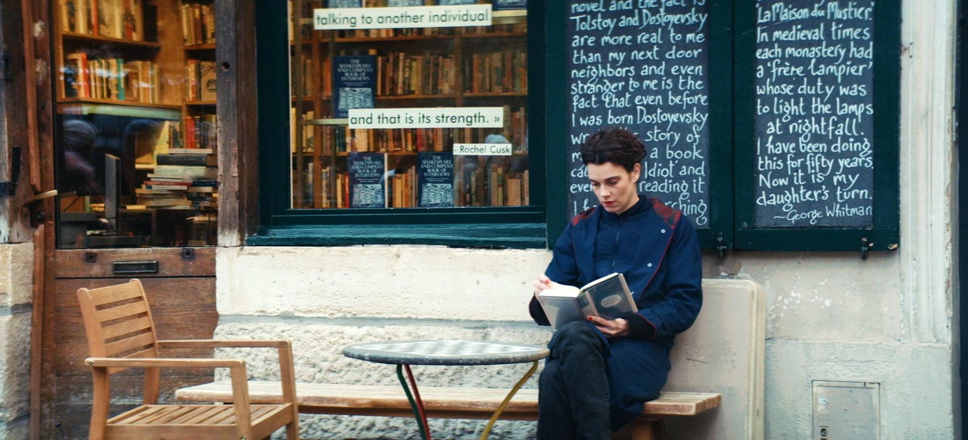 Eine Person liest ein Buch vor einem Buchladen. Bücherregale sind im Inneren sichtbar. Ein Tisch und ein Stuhl befinden sich im Vordergrund.