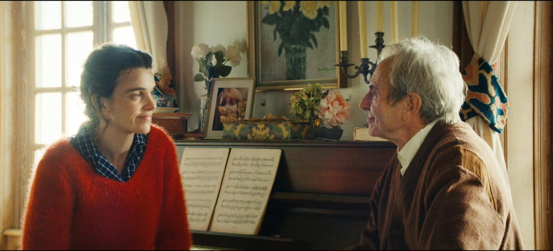 Eine Frau in einem roten Pullover und ein älterer Mann stehen sich in der Nähe eines Klaviers gegenüber, Notenblätter sichtbar. Blumen und ein Gemälde sind im Hintergrund.