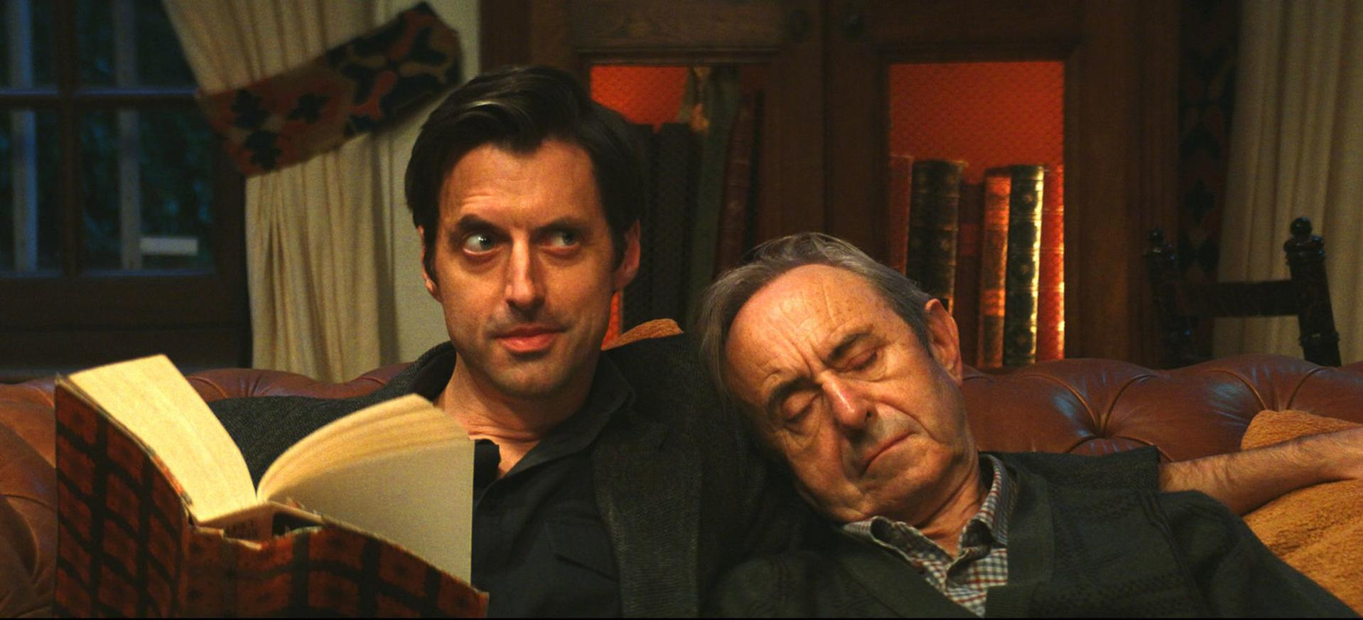 Zwei Männer auf einem Ledersofa. Einer liest ein Buch, der andere schläft. Bücherregale und ein Fenster im Hintergrund.