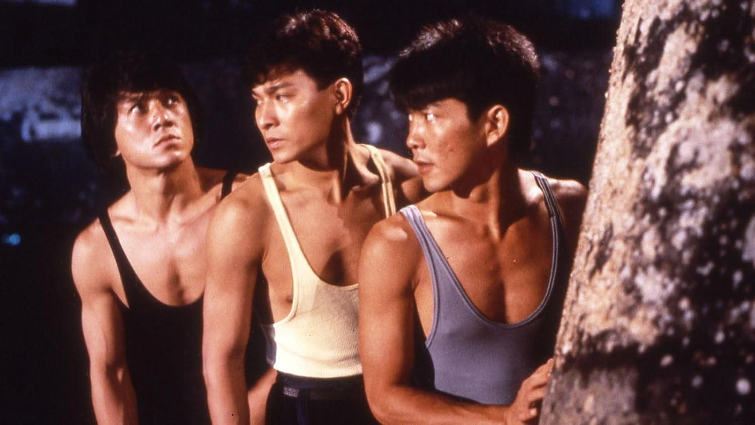 Drei Männer in Tanktops. Einer schaut nach oben, der zweite nach links, der dritte nach rechts, teilweise hinter einem Baum versteckt.