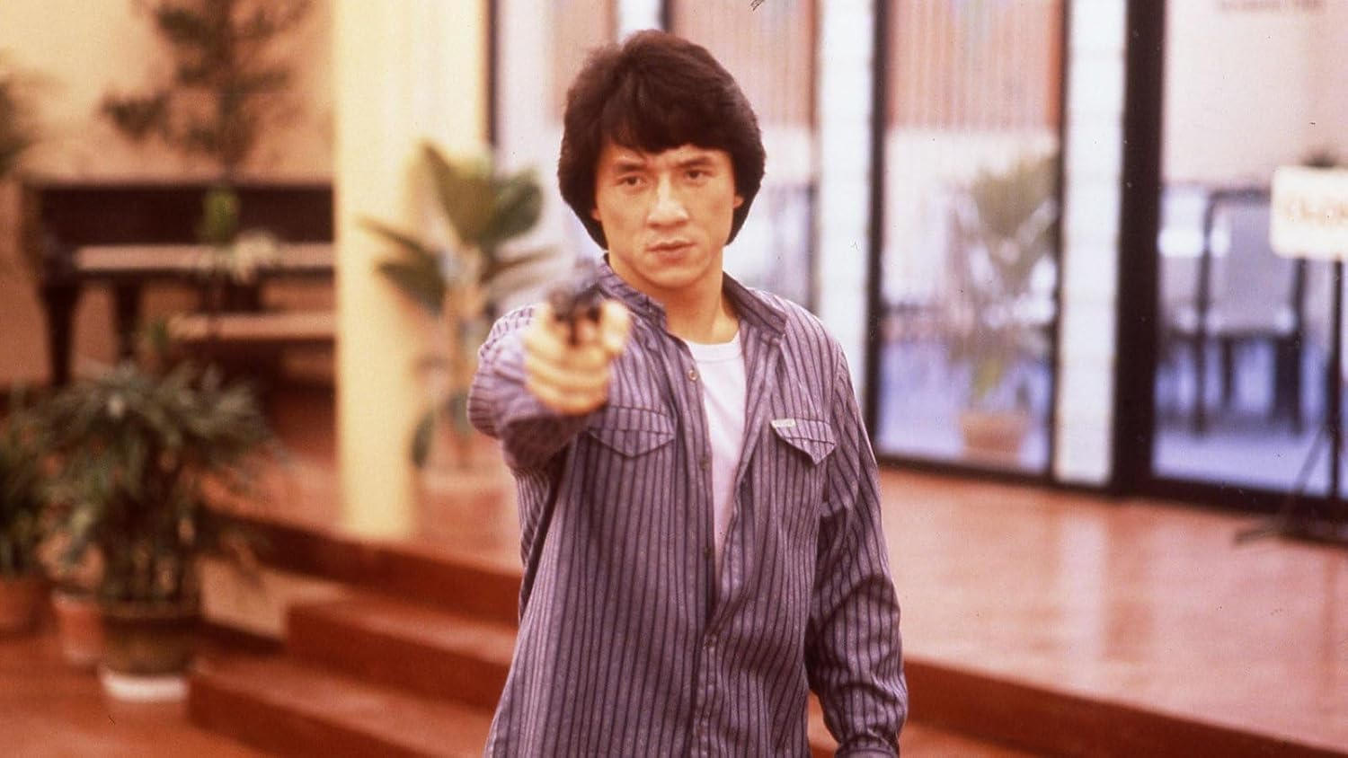 Jackie Chan zielt mit einer Waffe, trägt ein gestreiftes Hemd. Dahinter ist ein Raum mit Pflanzen und einem Klavier zu sehen.