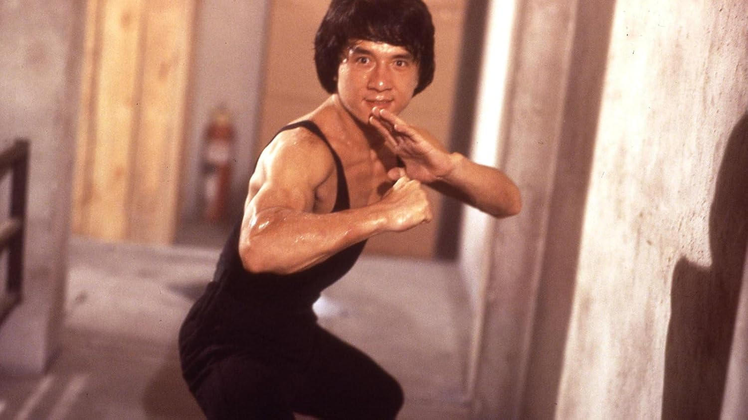 Jackie Chan in schwarzem Outfit, mit Fäusten, in Kampfhaltung. In einem Raum, Feuerlöscher sichtbar.