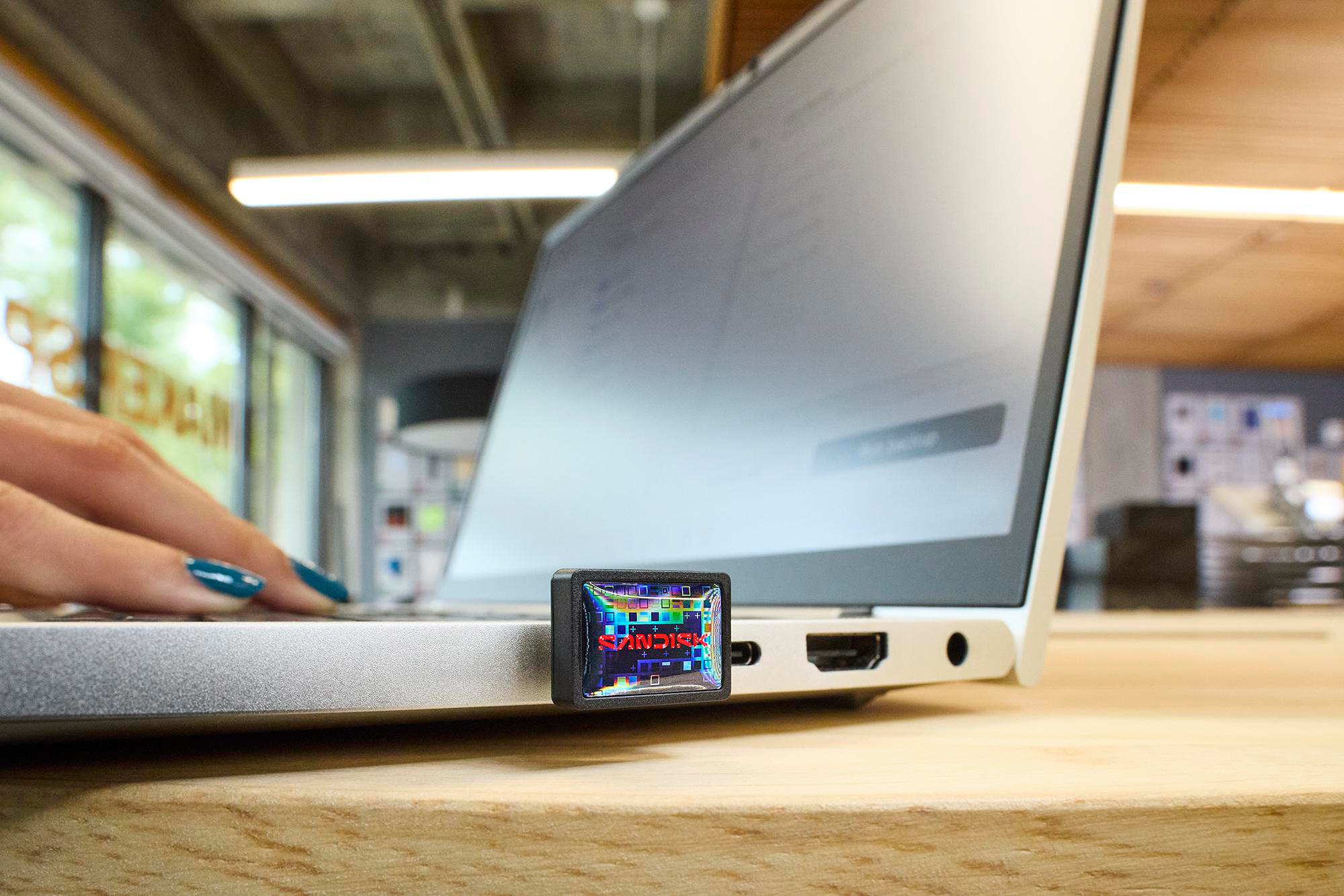 Ein Laptop mit einem SanDisk USB-Laufwerk. Eine Hand mit tealfarbenen Nägeln liegt auf dem Laptop. Ein Holztisch ist im Vordergrund.