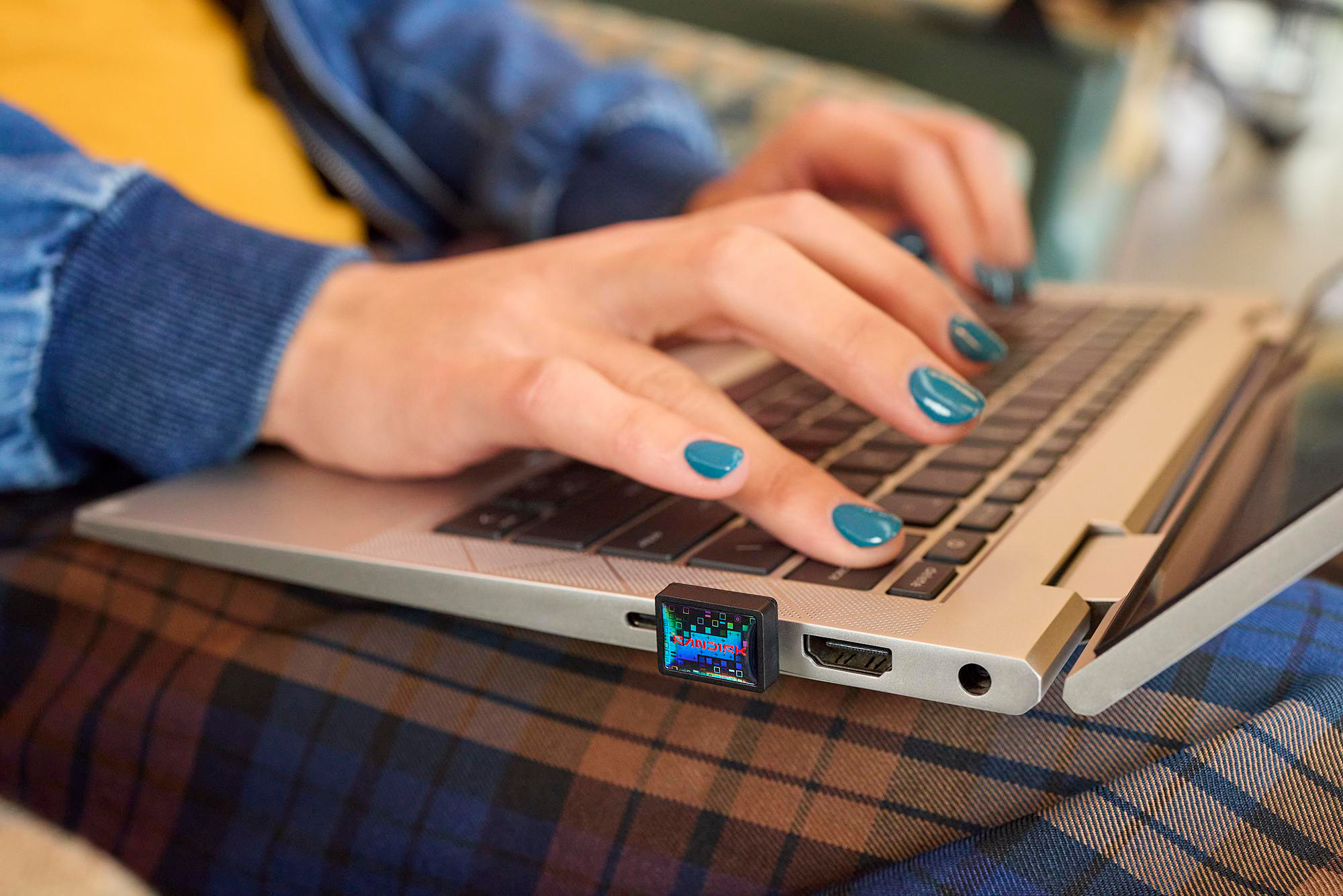 Eine Person tippt auf einem Laptop mit türkisfarbenen Fingernägeln. Ein kleines Gerät ist eingesteckt. Die Person trägt eine blaue Jacke und karierte Hose.