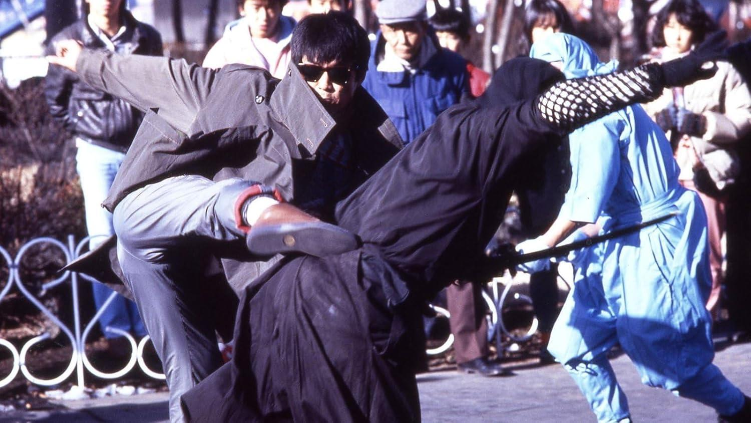 Ein Mann mit Sonnenbrille tritt einen anderen. Ein Ninja in Blau steht mit einem Schwert. Zuschauer beobachten den Kampf im Hintergrund.
