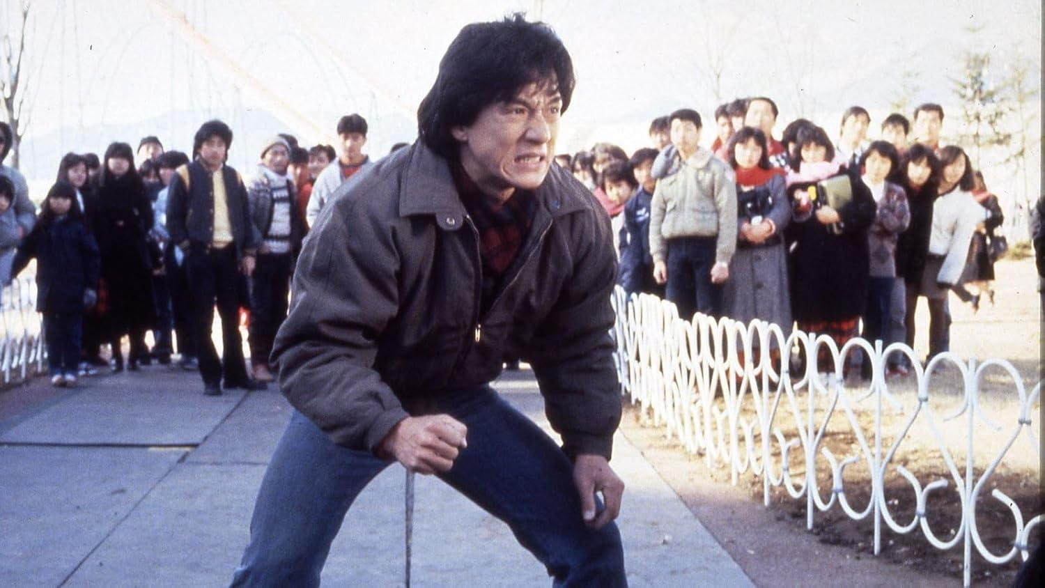 Jackie Chan in Kampfpose, umgeben von Menschen. Er trägt eine Jacke, Jeans und sieht wütend aus. Weißer Zaun und Menschenmenge im Hintergrund.