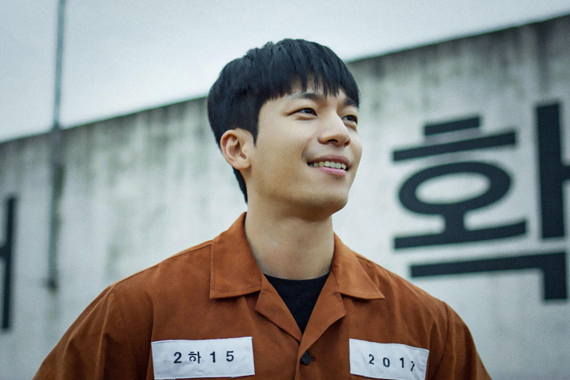 Ein lächelnder Mann in orangefarbener Gefängnisuniform steht vor einer Wand mit koreanischer Schrift. Aufnäher lesen '2 하 15' und '2017'.