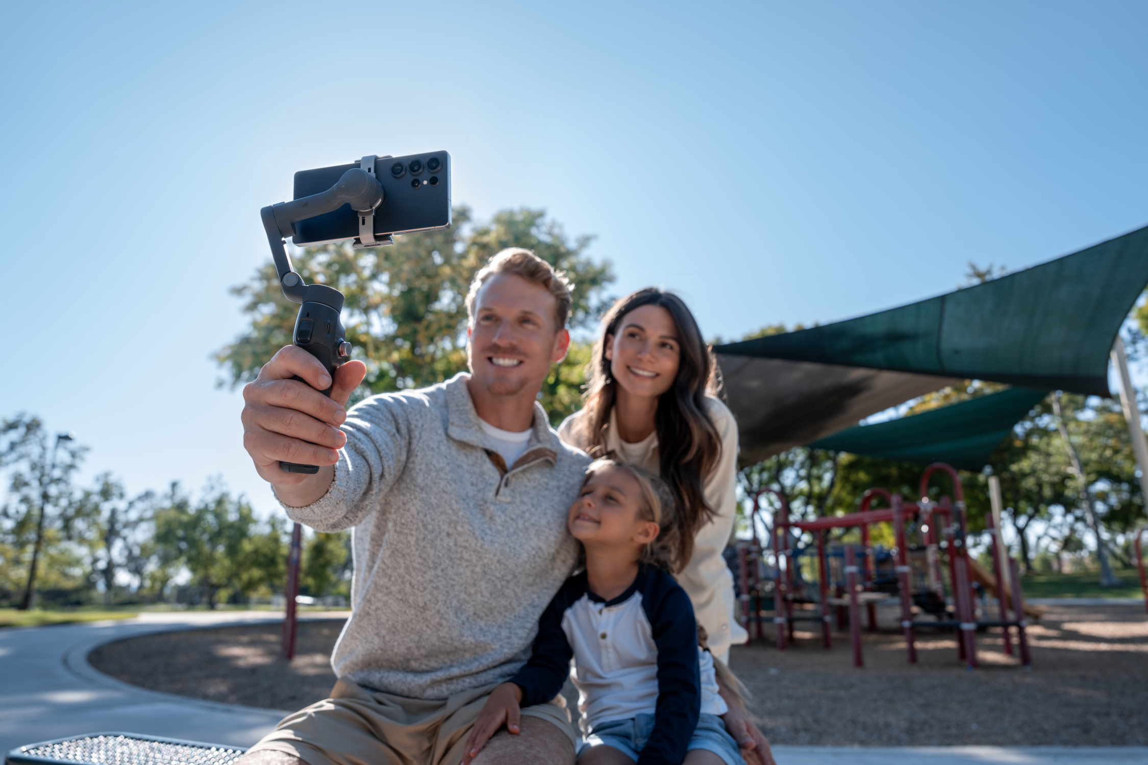 Una famiglia fa un selfie con un telefono su un gimbal. Sono in un parco con un parco giochi sullo sfondo sotto un cielo blu.