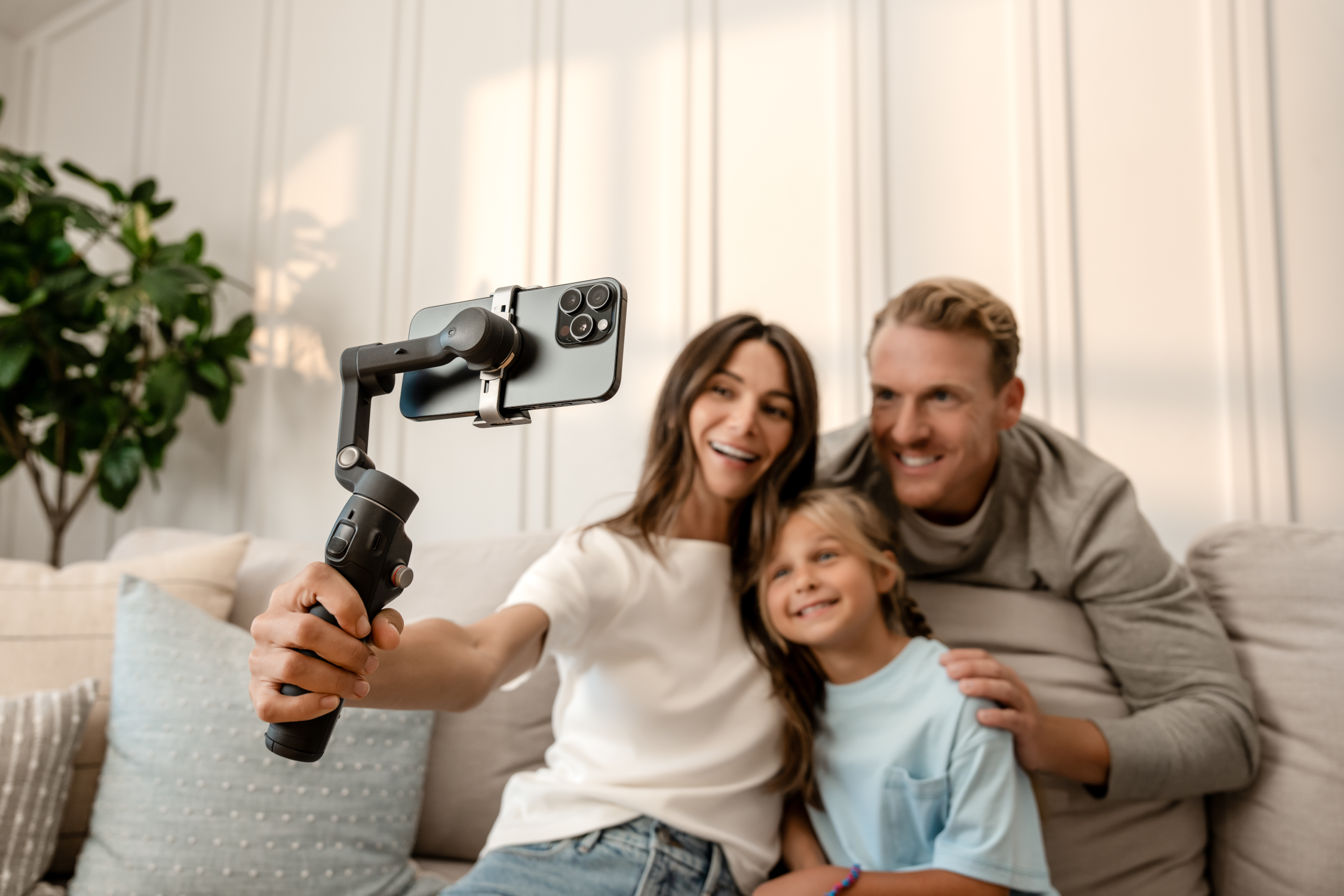 Una famiglia di tre persone si fa un selfie con uno smartphone. Sono su un divano. Lo sfondo ha una pianta e un muro bianco.