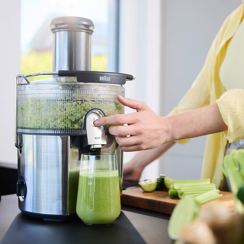 Una donna aziona un estrattore di succo, facendo succo verde. Un bicchiere di succo è sotto. Sedano e un lime sono su un tagliere.